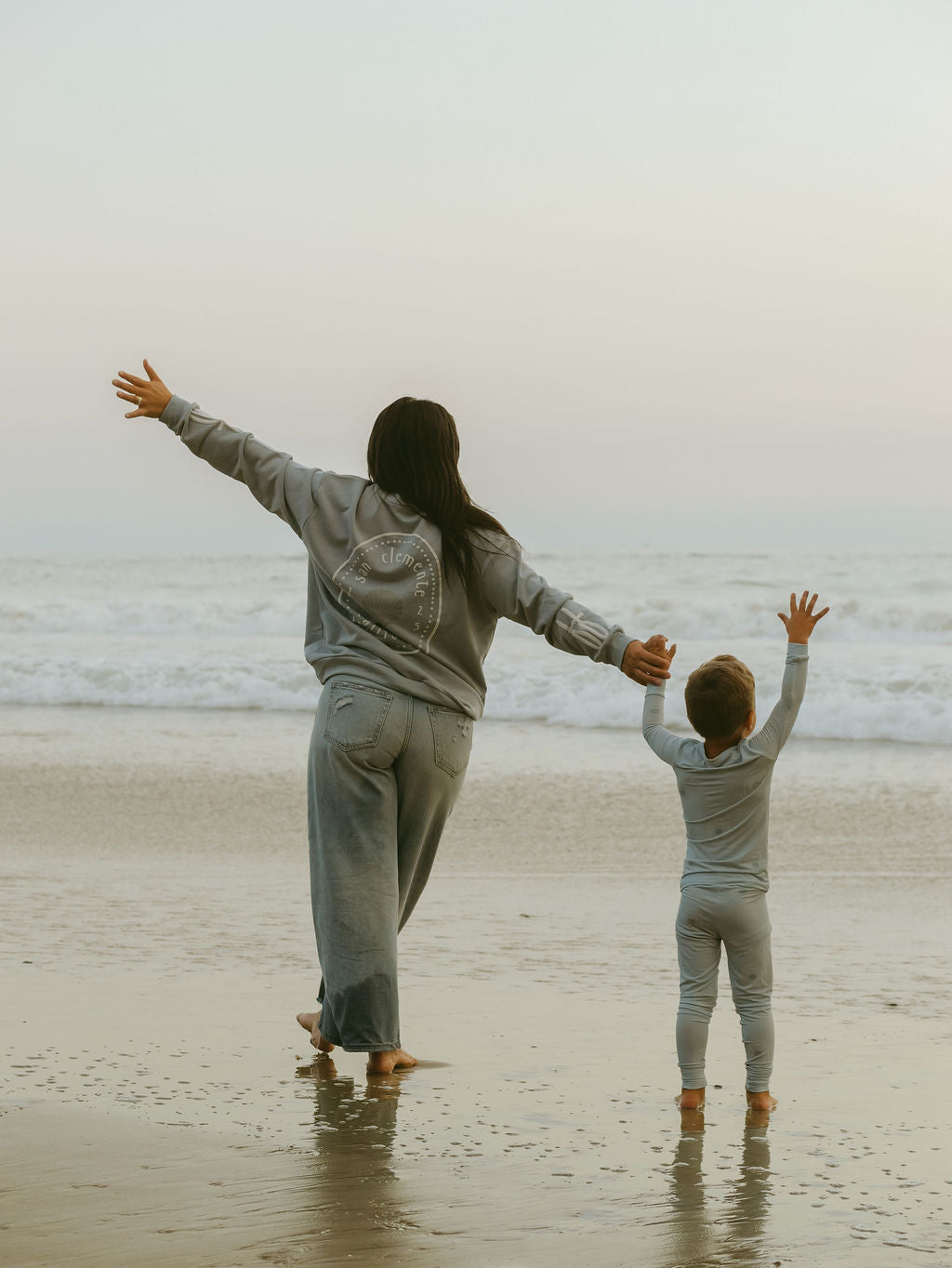 An adult and child in forever french baby's Bamboo Two Piece Pajamas | San Clemente stand barefoot on a beach with arms joyfully raised, overcast sky above and gentle waves rolling in behind them.