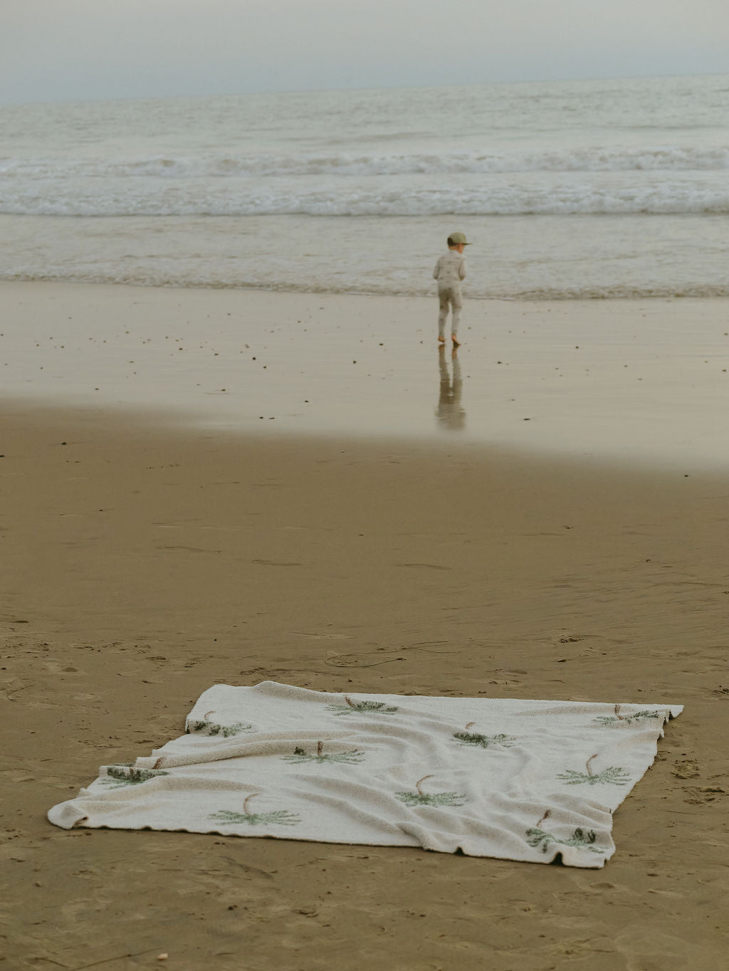 A forever french baby Plush Blanket | Palm Tree with a green design is laid on the sandy beach in the foreground as a child walks by the shoreline, facing the ocean waves.