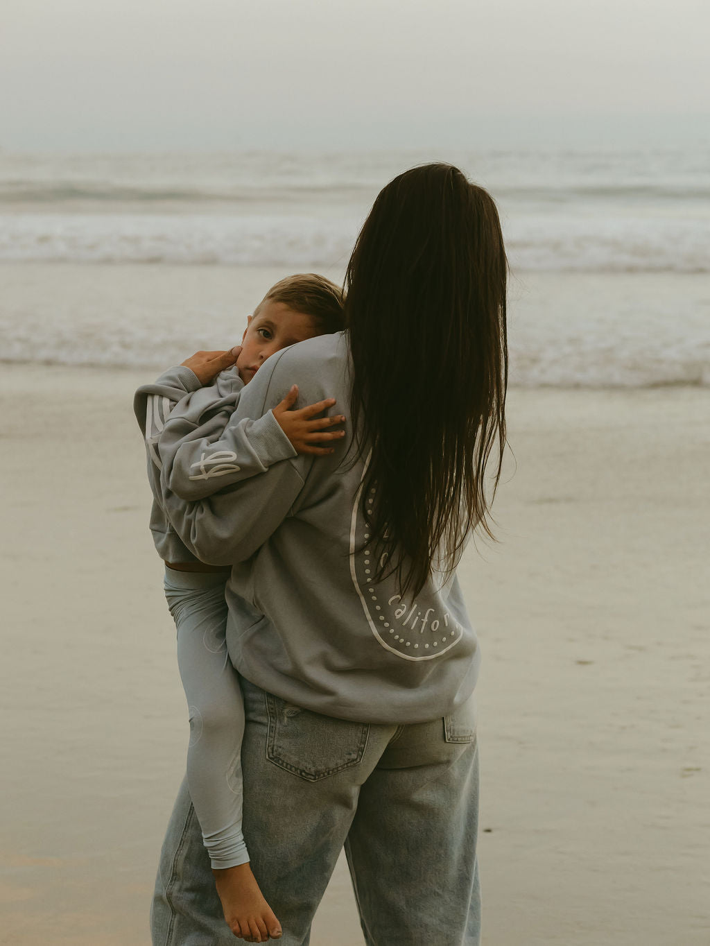 A woman stands on the beach holding a child who is wearing the forever french baby Child Sweatshirt | San Clemente, with ocean waves in the background, creating a perfect coastal keepsake.