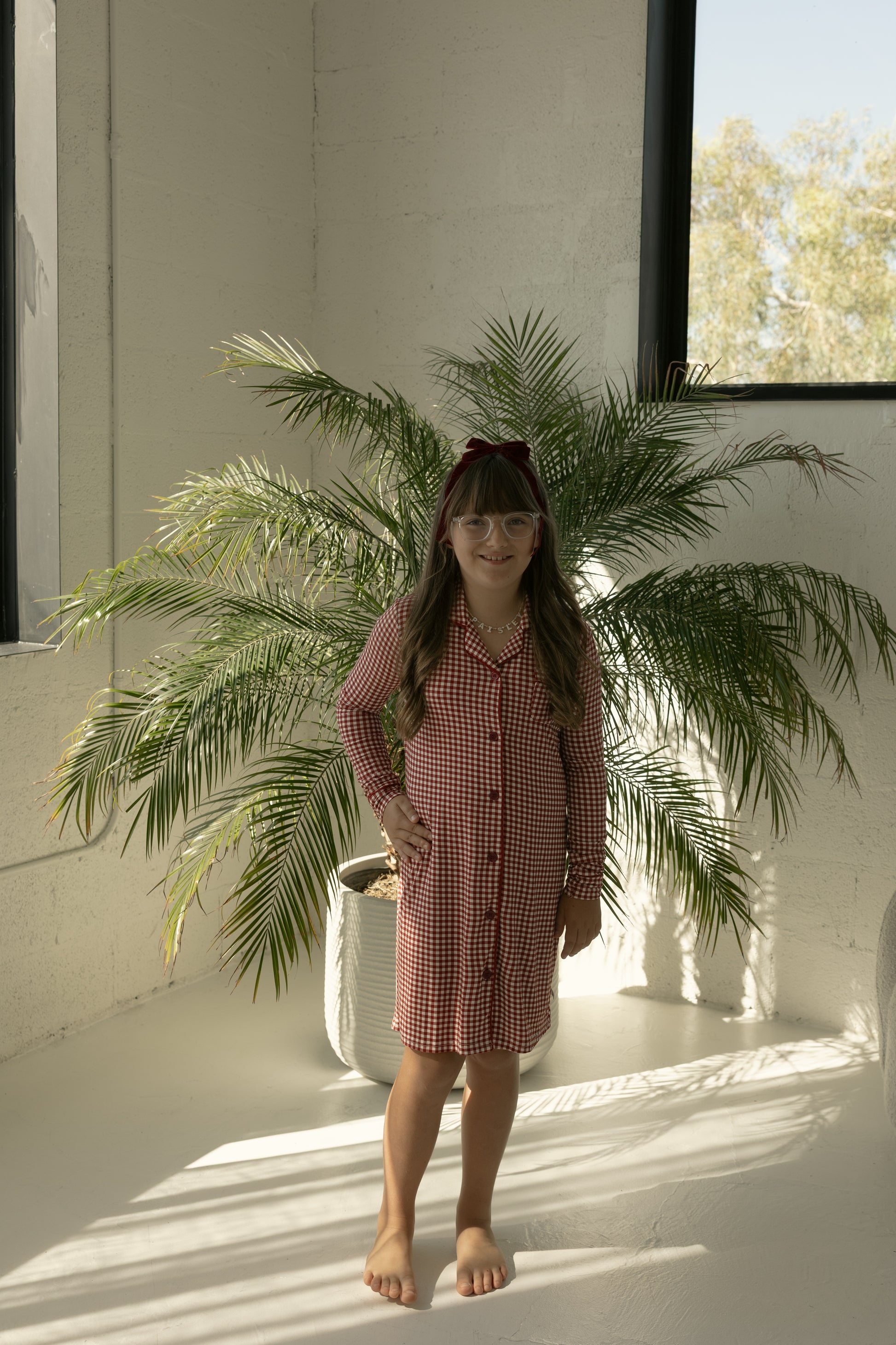 A young girl in the forever french baby Toddler Bamboo Sleeping Dress | Red Gingham stands barefoot by a sunlit window near a potted palm, sunlight highlighting her cozy red gingham outfit.
