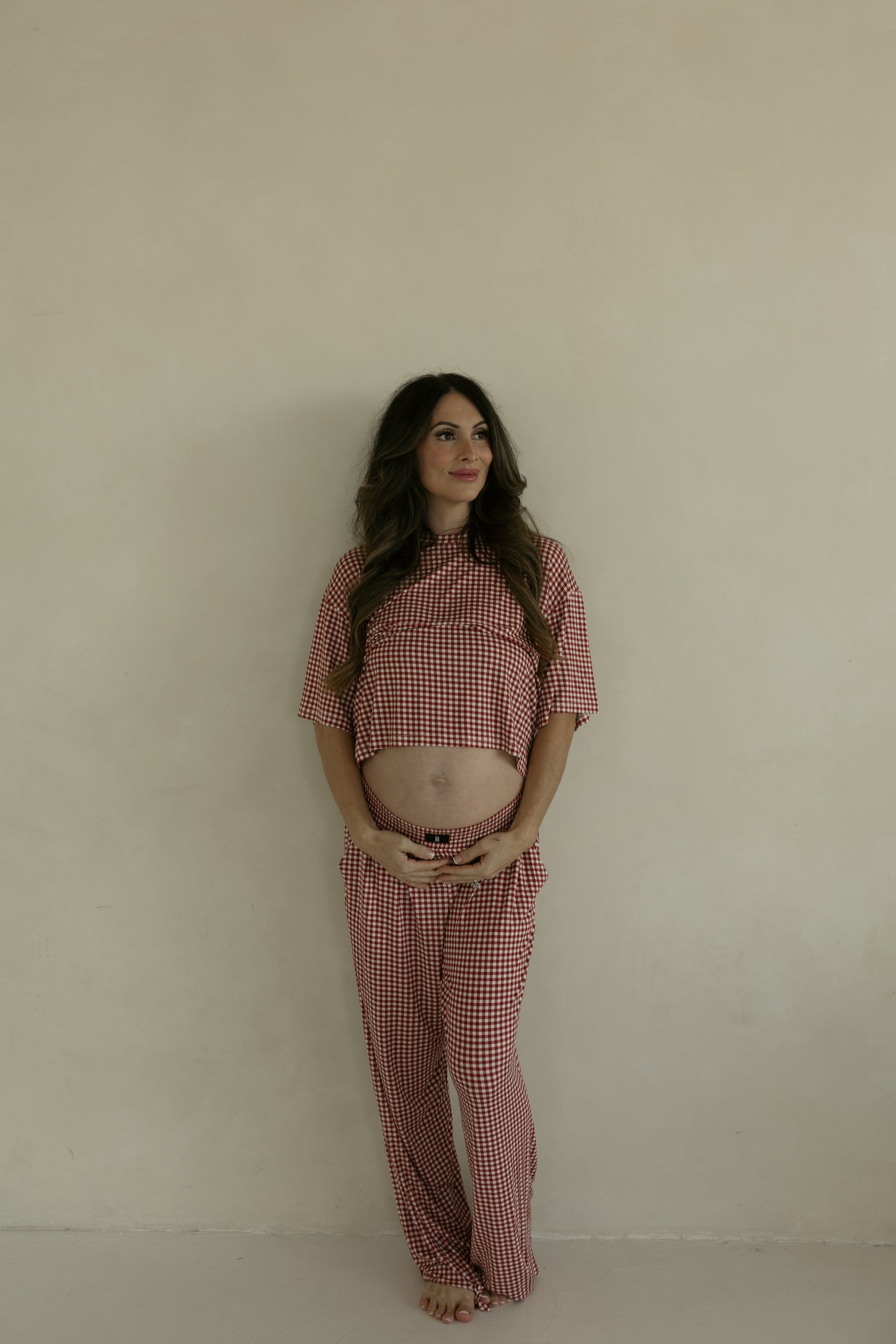 A pregnant woman with long brown hair stands barefoot against a beige wall, smiling softly as she wears the forever french baby Women's Boxy Tee Lounge Set in Red Gingham, her shirt lifted to show her baby bump and radiating comfort.