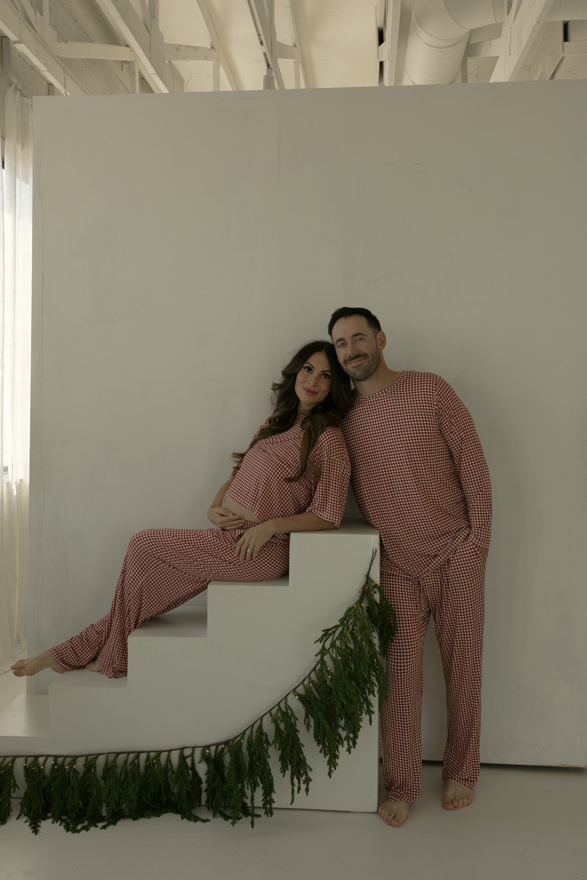 A couple in coordinated forever french baby loungewear poses indoors; the pregnant woman wears the Women's Boxy Tee Lounge Set in Red Gingham as they smile together on white steps decorated with greenery.