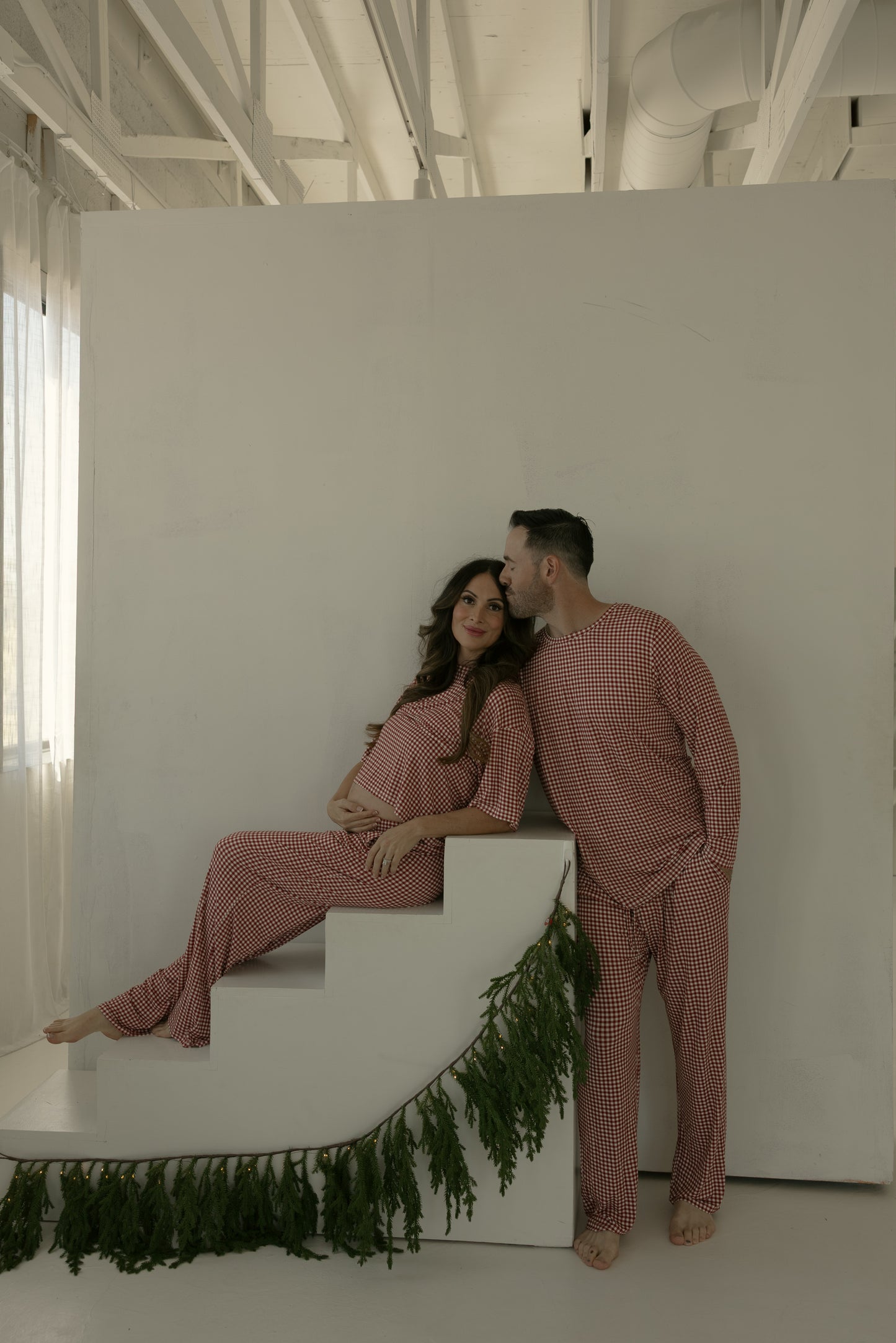 A woman wears the forever french baby Women's Boxy Tee Lounge Set in Red Gingham as she sits on white stairs with greenery; a man in matching pajamas stands nearby, kissing her head. The softly lit scene features white walls and exposed ceilings.