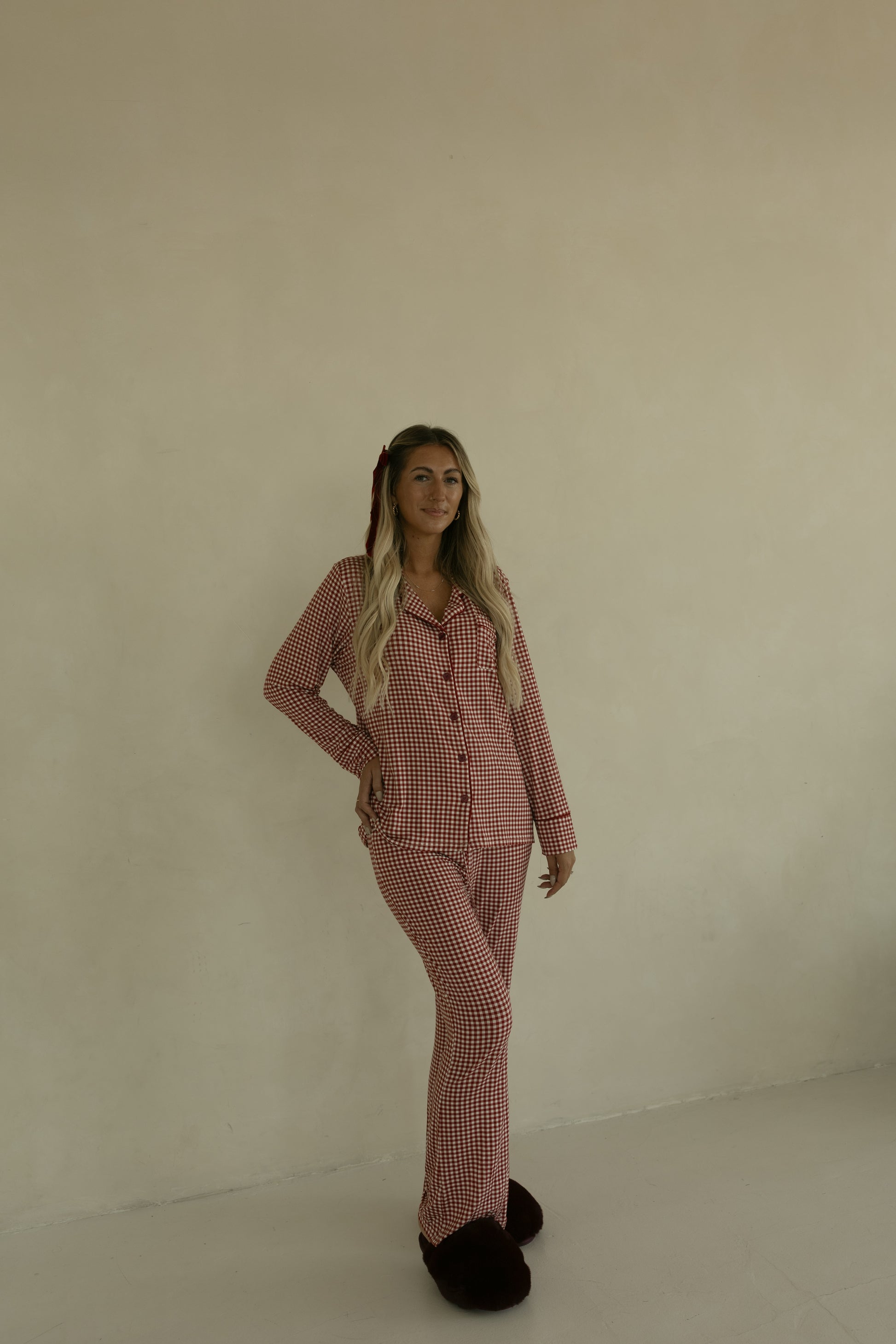 A woman with long blonde hair, smiling slightly with one hand on her hip, stands against a beige wall wearing forever french baby’s Long Sleeve Women's Bamboo Pajamas in Red Gingham and dark slippers.