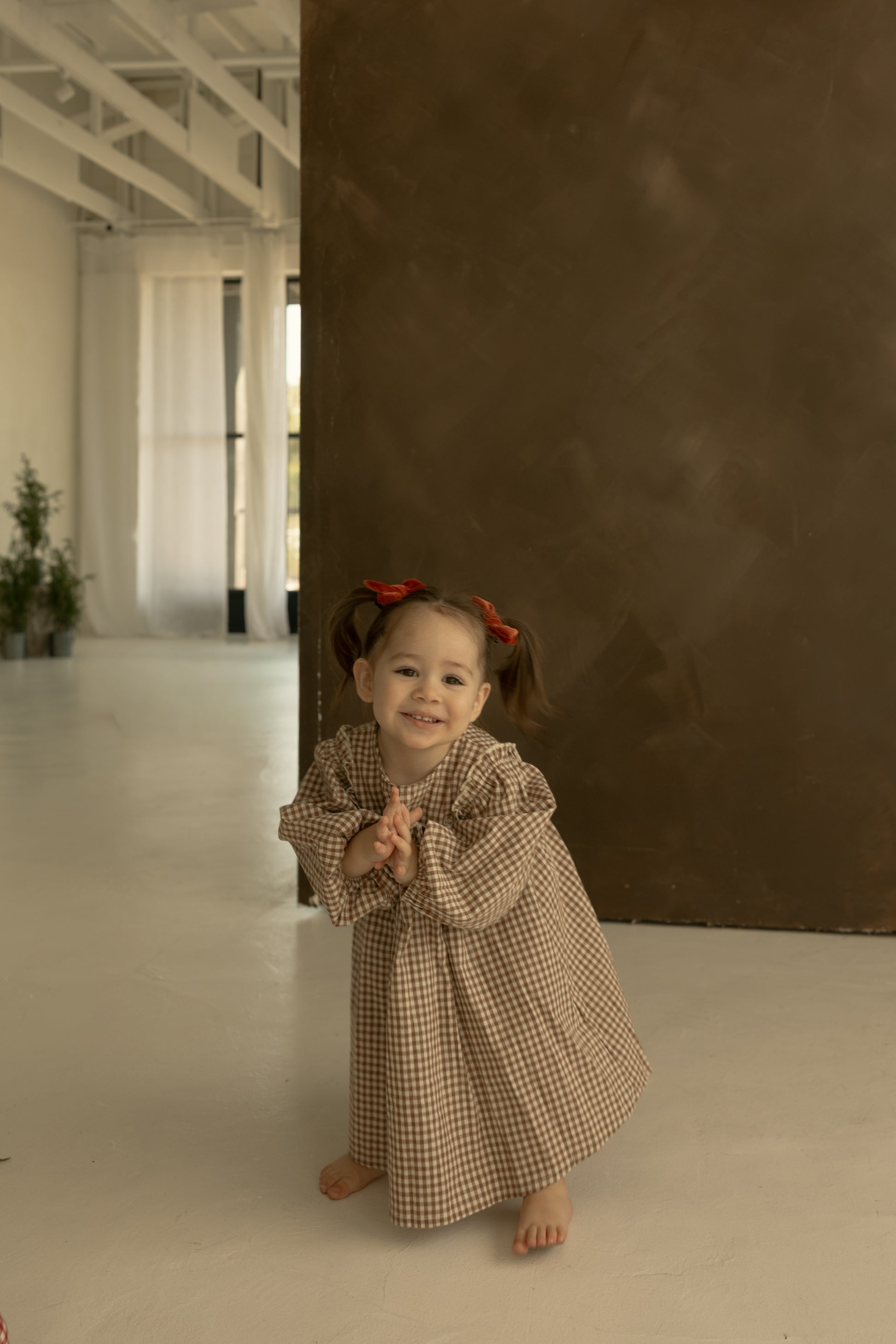 A young girl with pigtails and red bows smiles barefoot, clasping her hands, in front of a brown wall and white curtains, wearing the forever french baby Child Long Sleeve Dress | Honey Gingham.