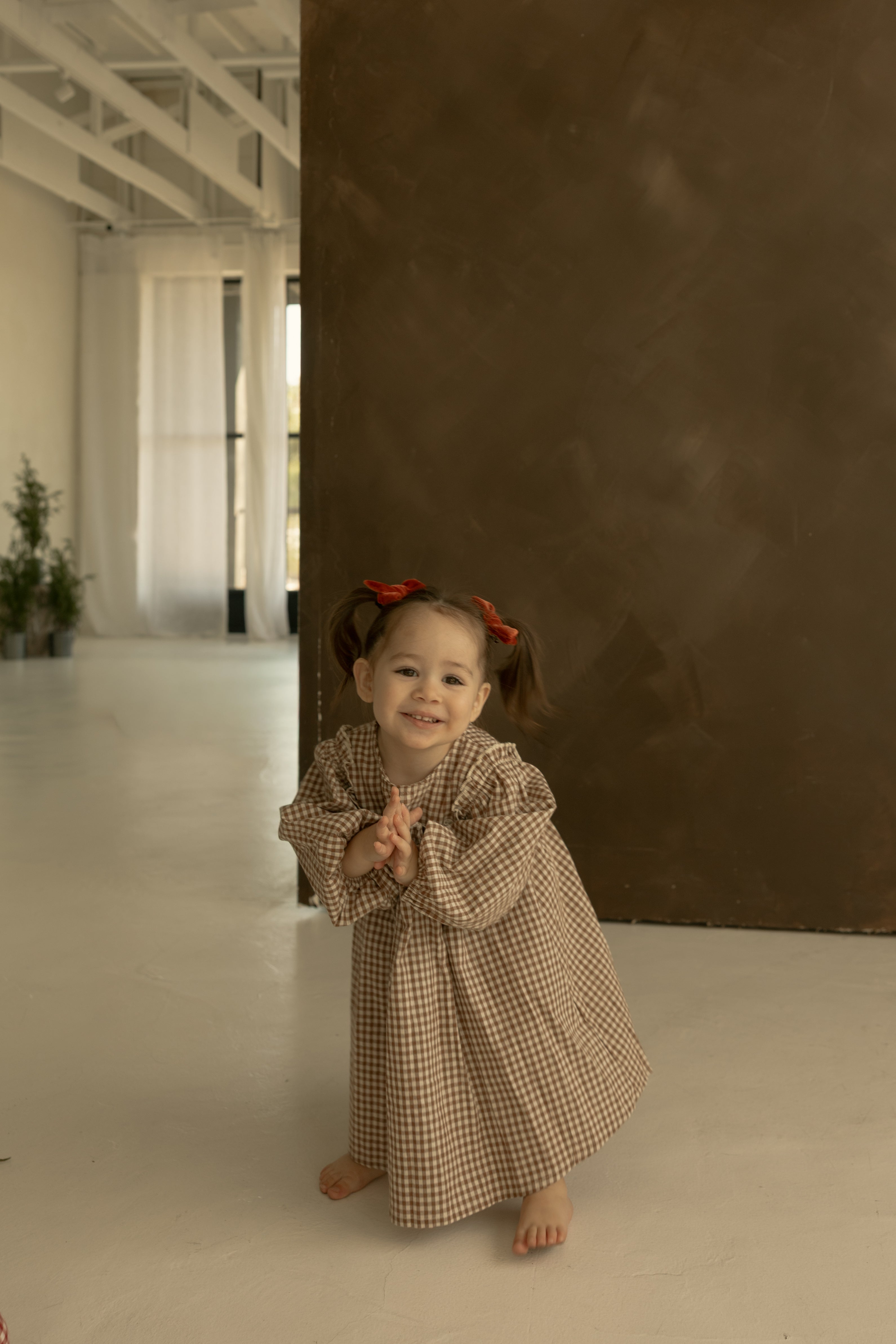 A young girl with pigtails and red bows smiles barefoot, clasping her hands, in front of a brown wall and white curtains, wearing the forever french baby Child Long Sleeve Dress | Honey Gingham.