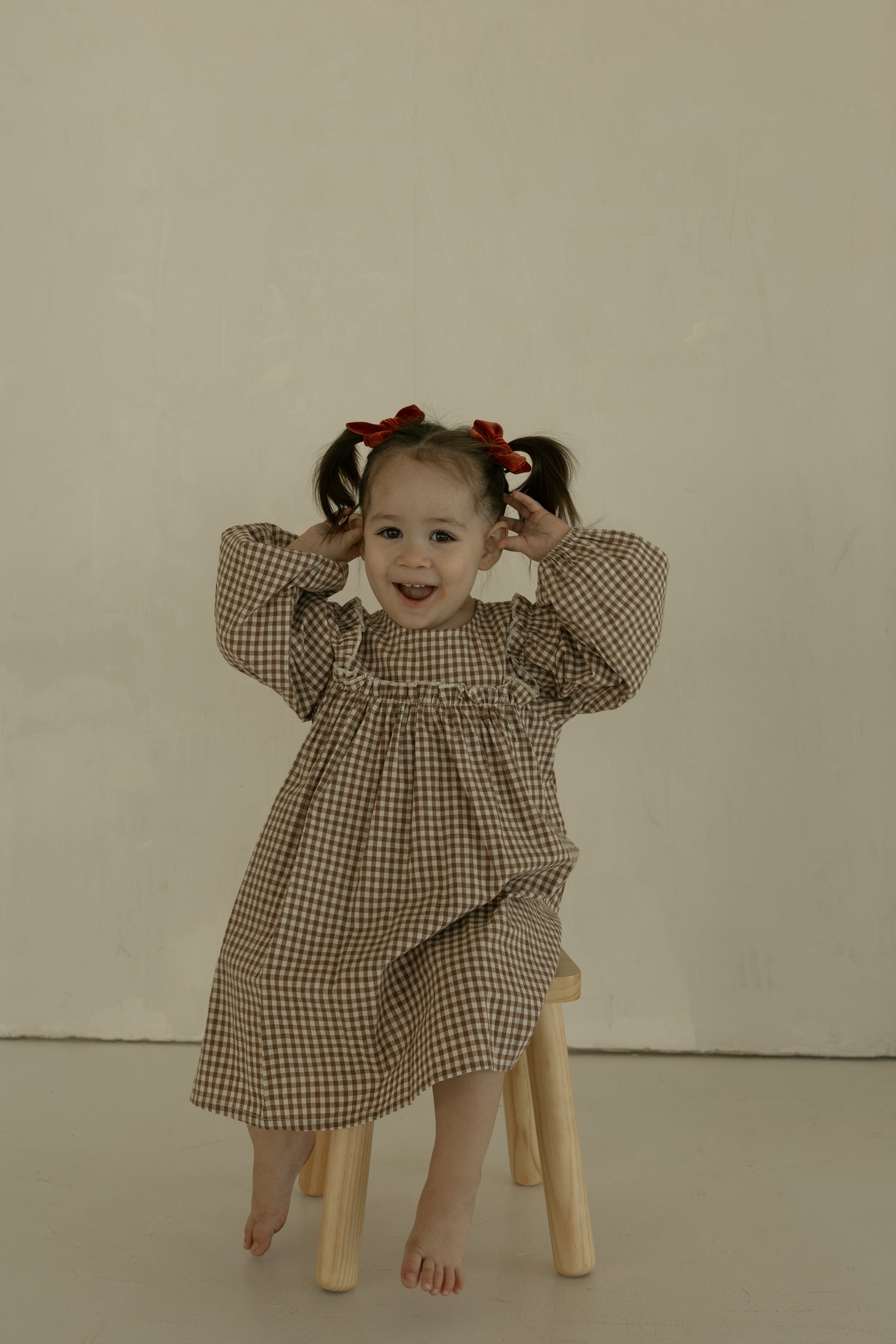 A young child with pigtails tied with red bows sits barefoot on a small wooden stool, smiling and holding her hands near her head, wearing the forever french baby Child Long Sleeve Dress | Honey Gingham against a plain background.