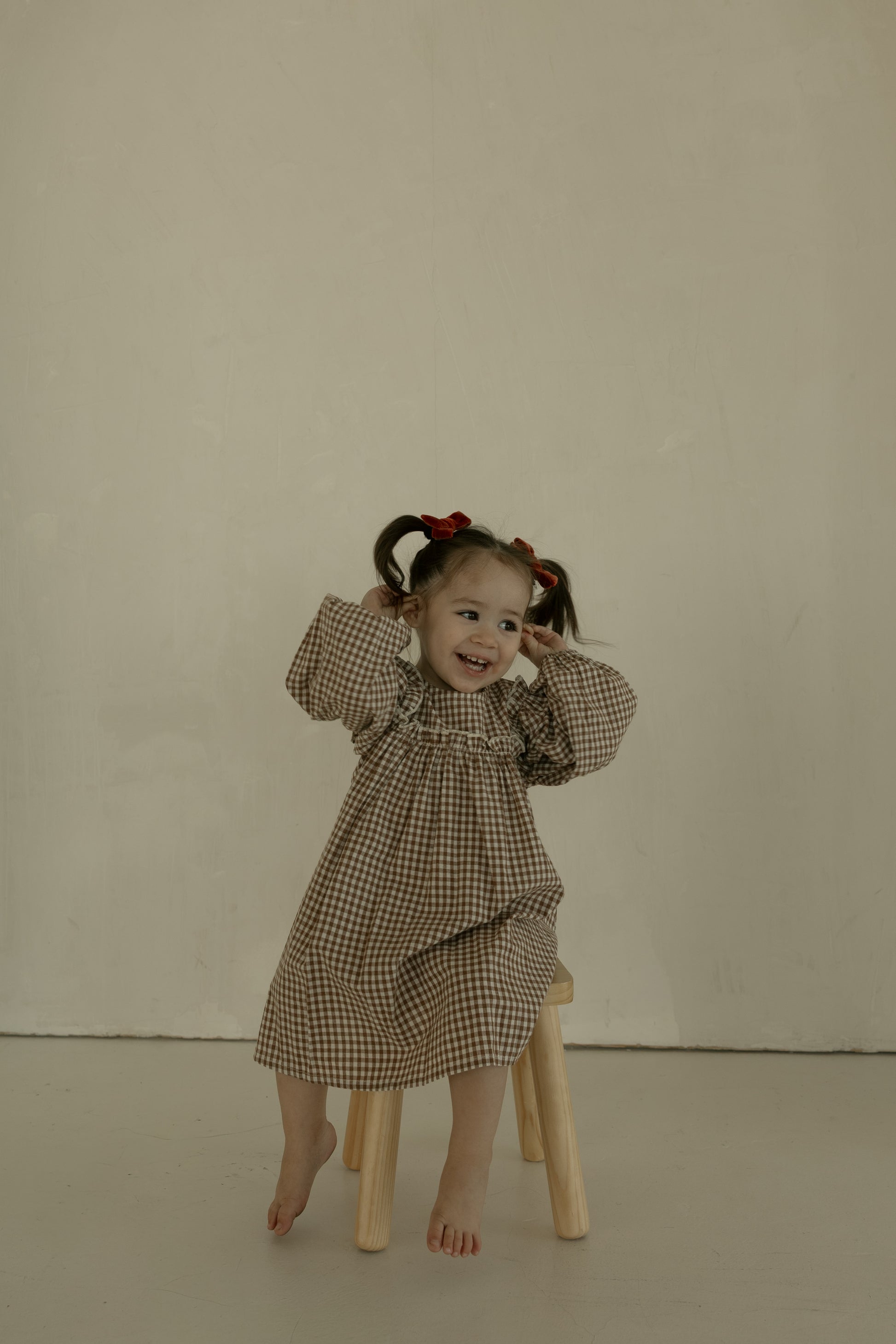 A young girl with pigtails and red bows sits barefoot on a stool, smiling and posing in the forever french baby Child Long Sleeve Dress | Honey Gingham against a plain, light background.