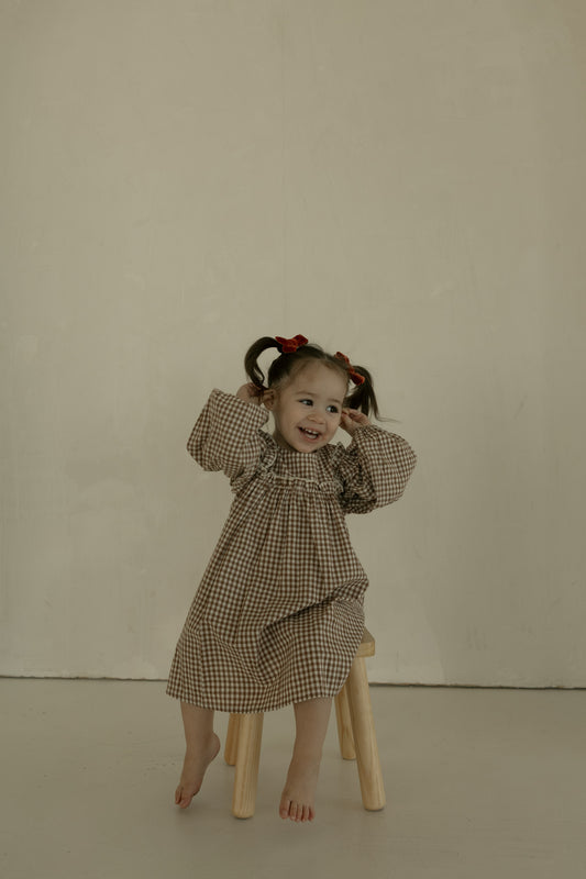 A young girl with pigtails and red bows sits barefoot on a stool, smiling and posing in the forever french baby Child Long Sleeve Dress | Honey Gingham against a plain, light background.