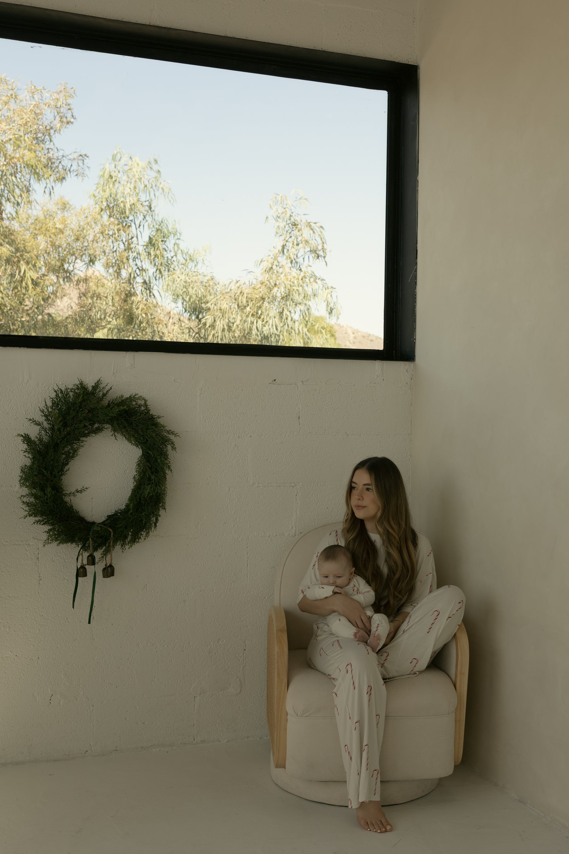 A woman in the Women's Boxy Tee Lounge Set | Candy Cane by forever french baby sits barefoot in a beige chair with a baby on her lap beside a green wreath and a window showing trees and blue sky.