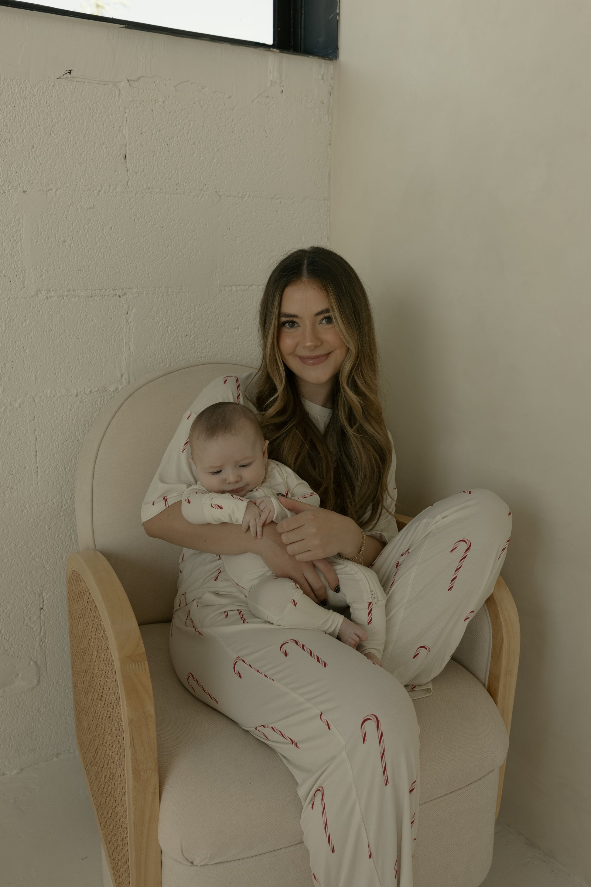 A woman with long hair smiles in a beige armchair, holding a baby. Both wear matching forever french baby Women's Boxy Tee Lounge Set | Candy Cane pajamas, featuring white fabric with red candy cane prints, against light-colored walls.