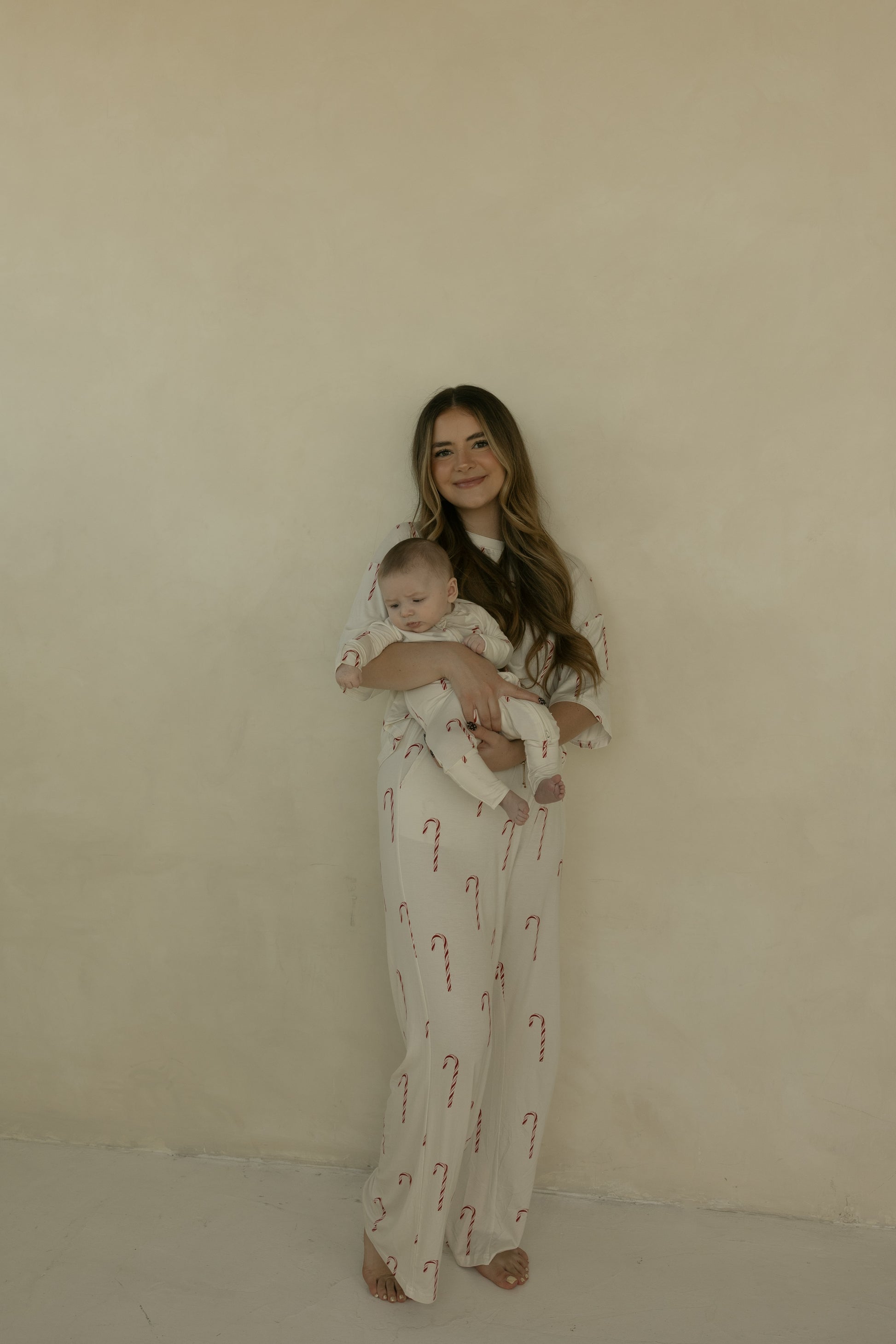 A woman with long hair stands barefoot against a beige wall, smiling and holding a baby. Both wear matching forever french baby Women's Boxy Tee Lounge Set | Candy Cane pajamas with red candy cane patterns.