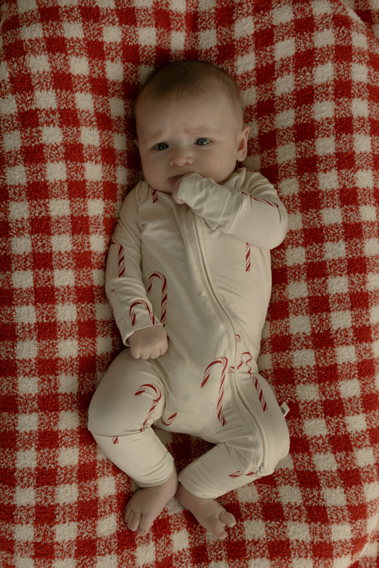 A baby in forever french baby's Bamboo Zip Pajamas | Candy Cane—a cream bamboo viscose onesie with red candy canes—lies on a red and white checkered blanket, hand near mouth, offering a hypo-allergenic sleepwear option.