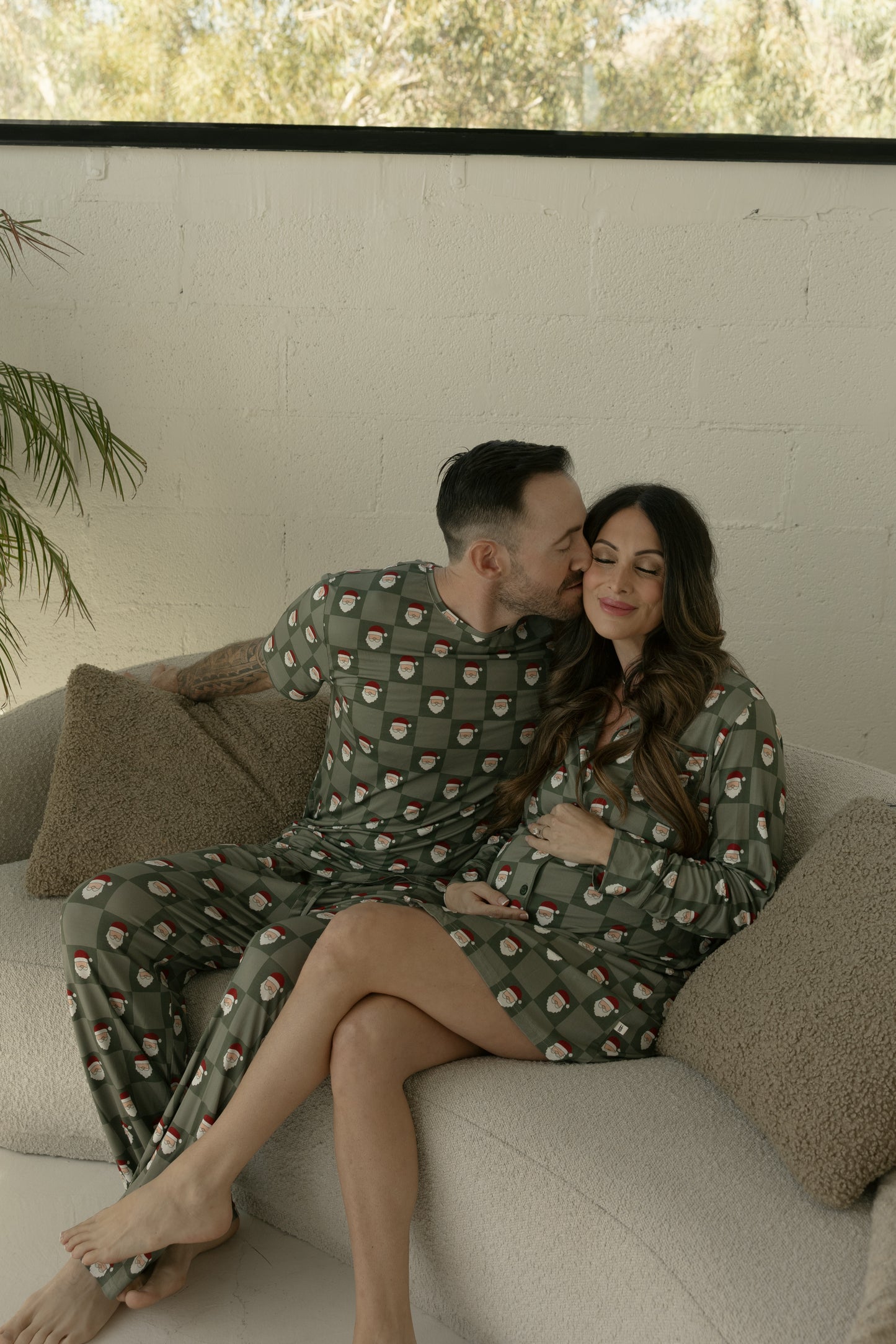 A pregnant woman in a forever french baby Women’s Bamboo Sleeping Dress | Santa Checkerboard smiles while holding her baby bump as she sits on a couch with her partner, both in matching hypoallergenic sleepwear.