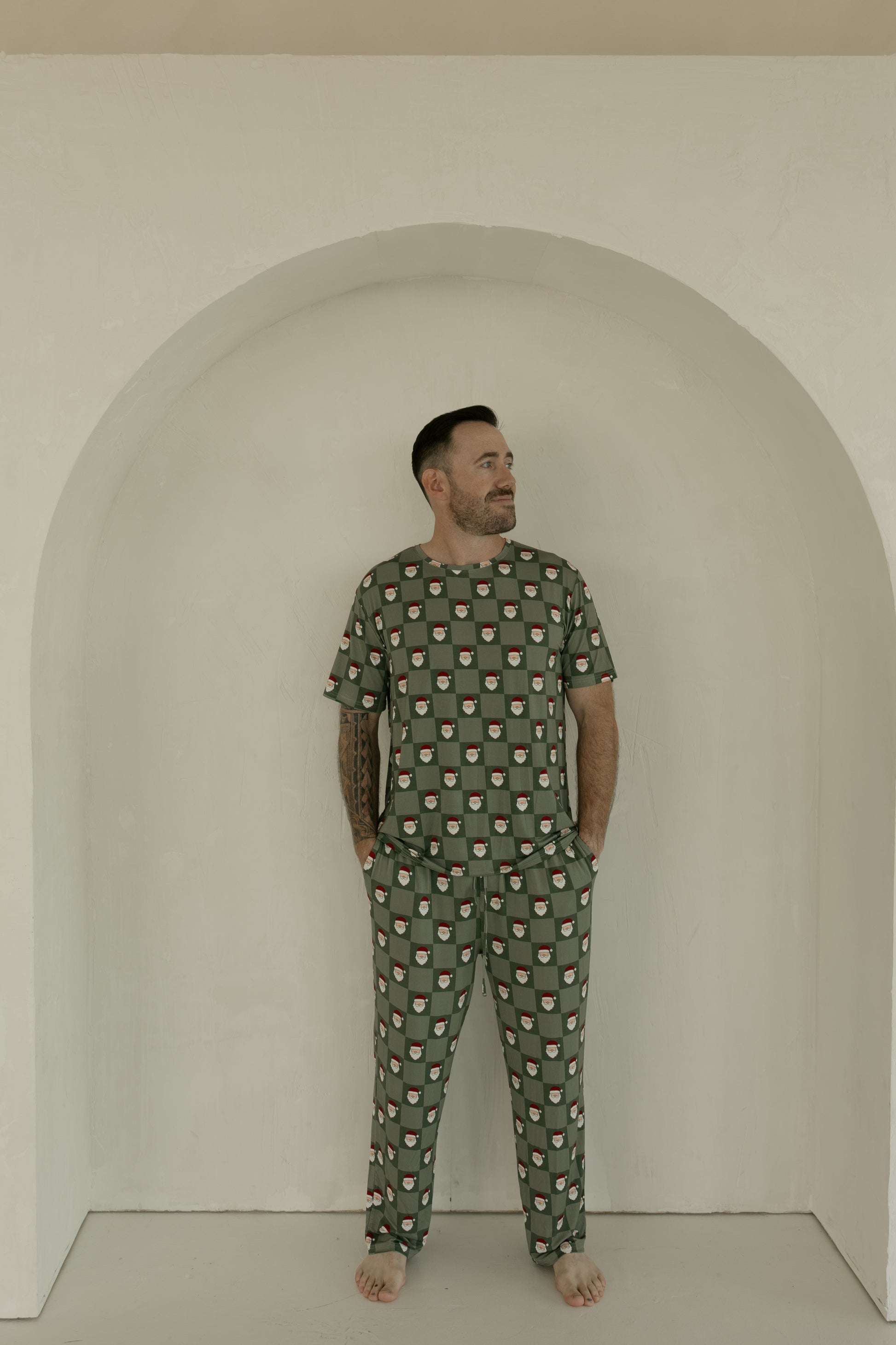 A bearded man stands barefoot in a white arched alcove, wearing forever french baby's Men's Short Sleeve Bamboo Pajamas in the Santa Checkerboard pattern. He has his hands in his pockets and looks slightly to his left.