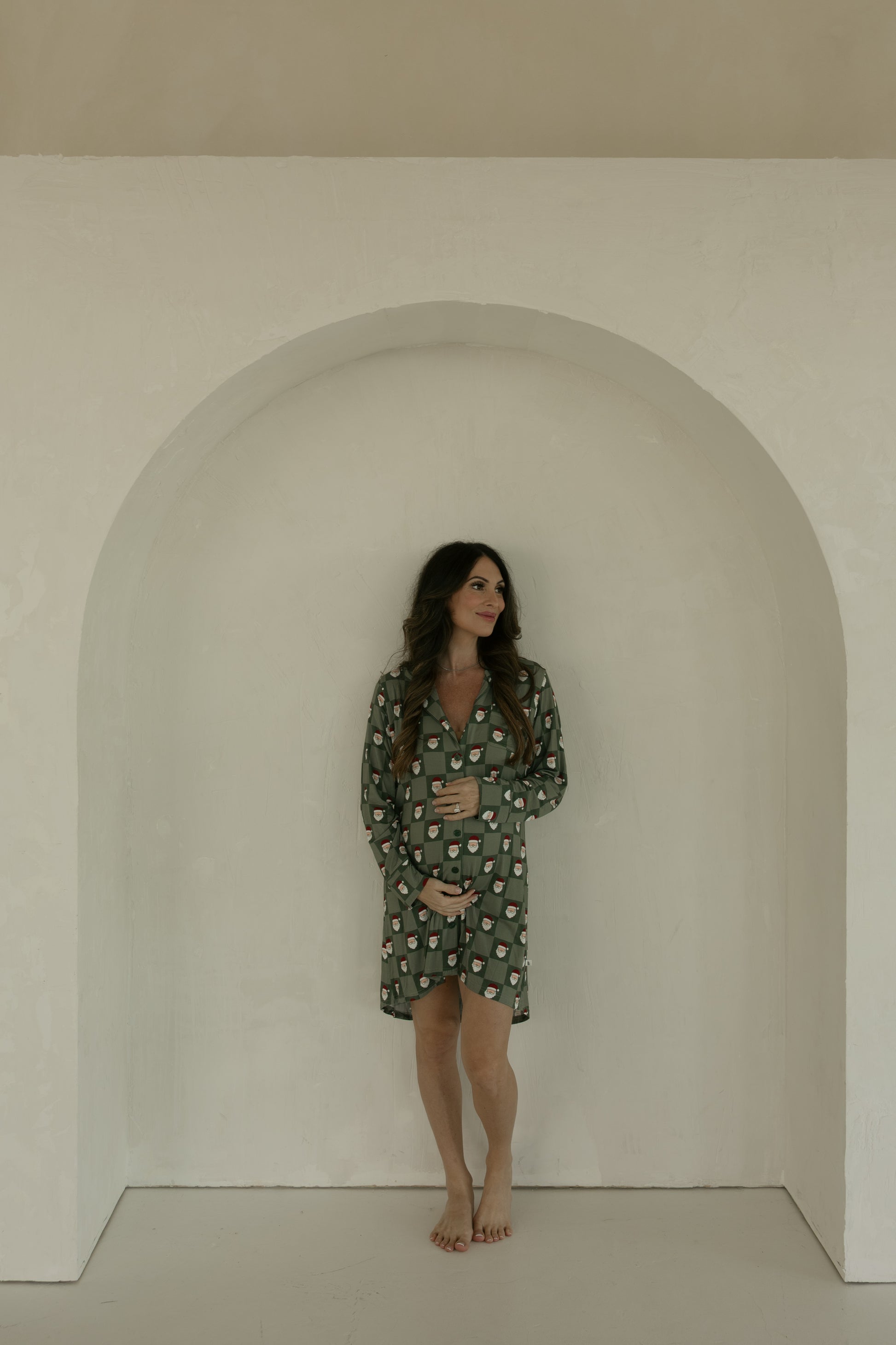 A woman with long brown hair stands barefoot in a light alcove, wearing the forever french baby Women's Bamboo Sleeping Dress in Santa Checkerboard, gently resting her hand on her belly and looking to the side.