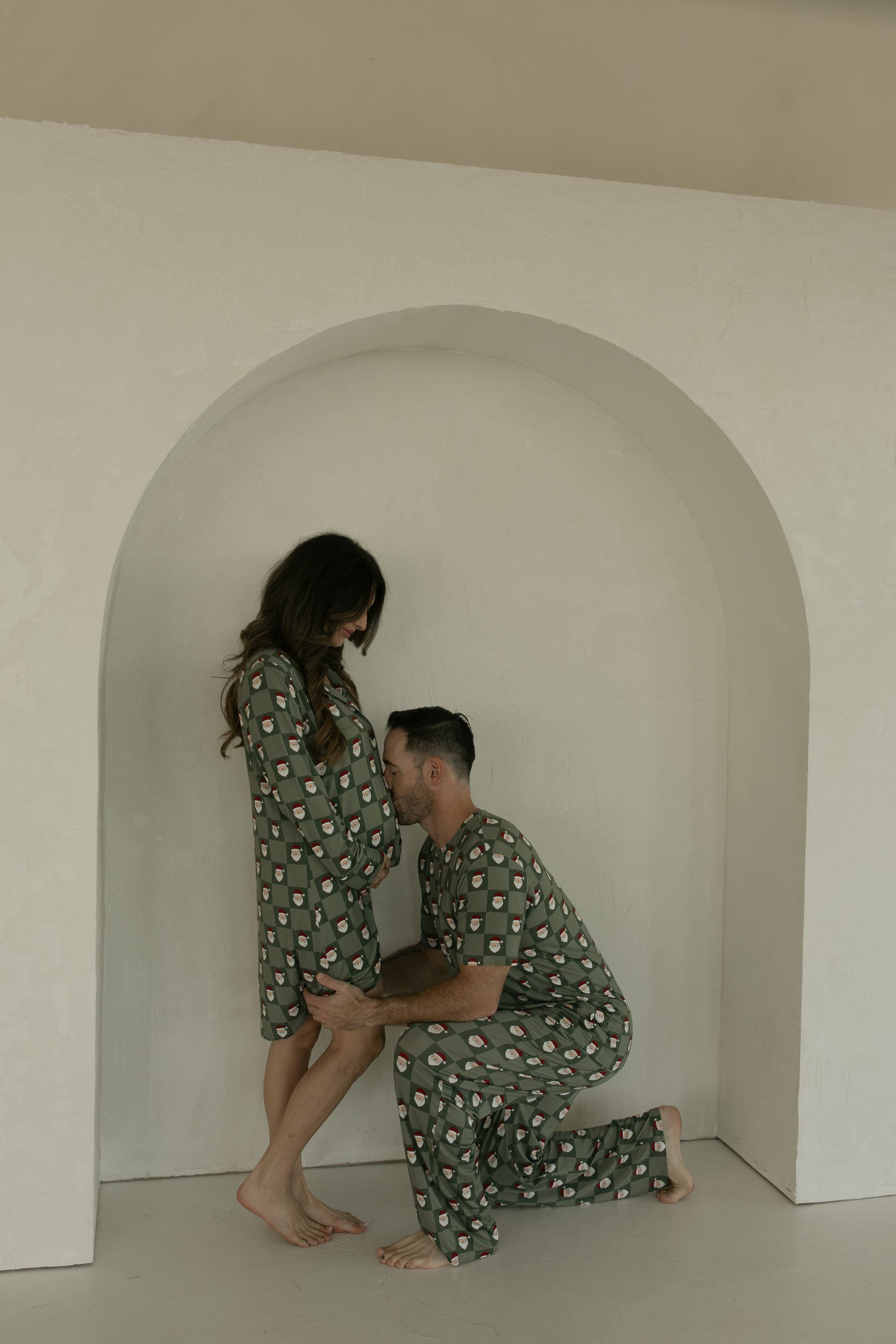 A man kneels and kisses a woman's pregnant belly as they wear matching green Santa Checkerboard sleepwear from forever french baby, with her in the Women's Bamboo Sleeping Dress, in front of a light arched alcove.