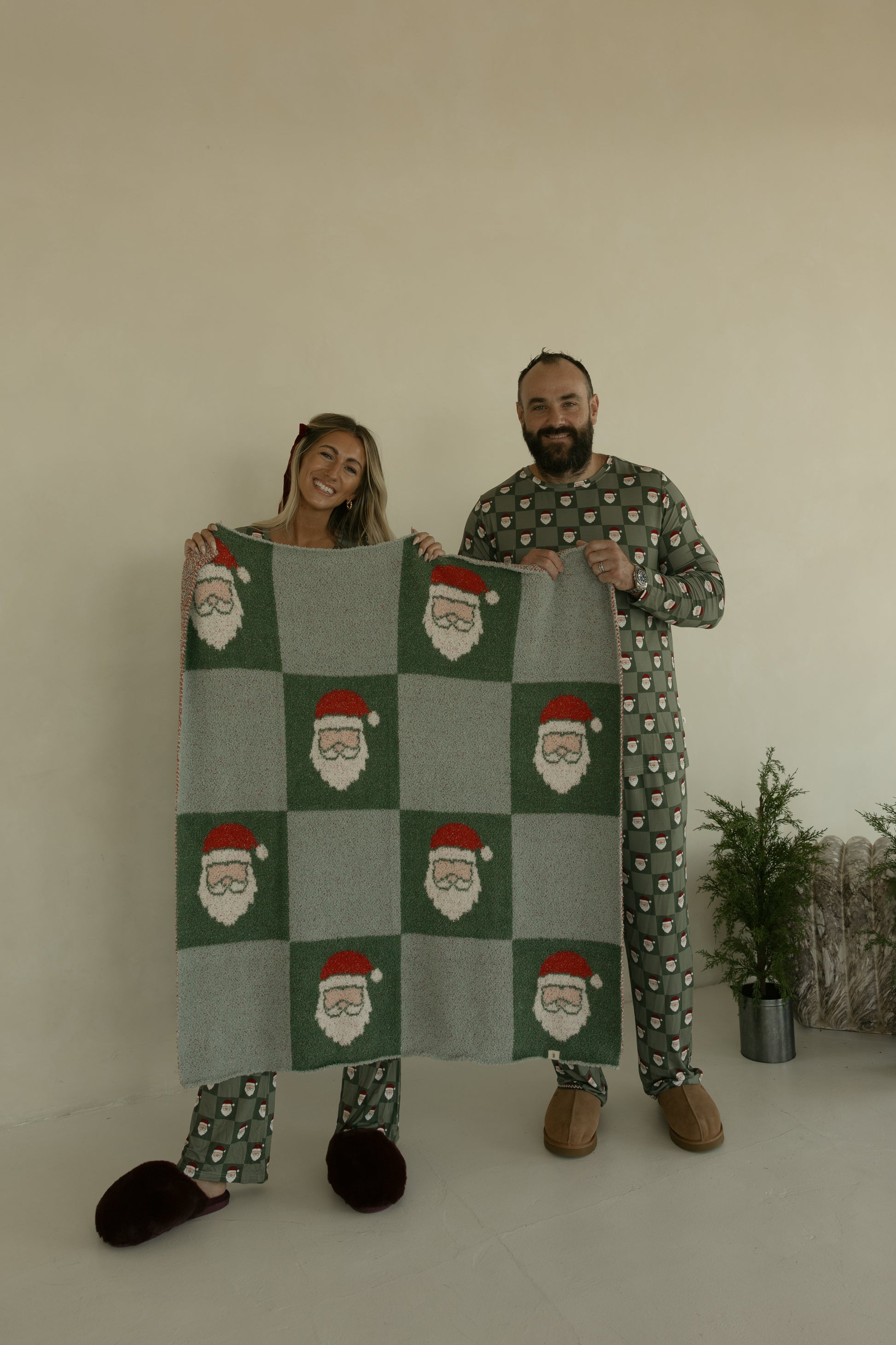 Two people in matching Christmas pajamas smile indoors as they hold up the forever french baby Plush Blanket | Santa Checkerboard, Oeko-Tex certified microfiber, beside two small potted evergreen trees.