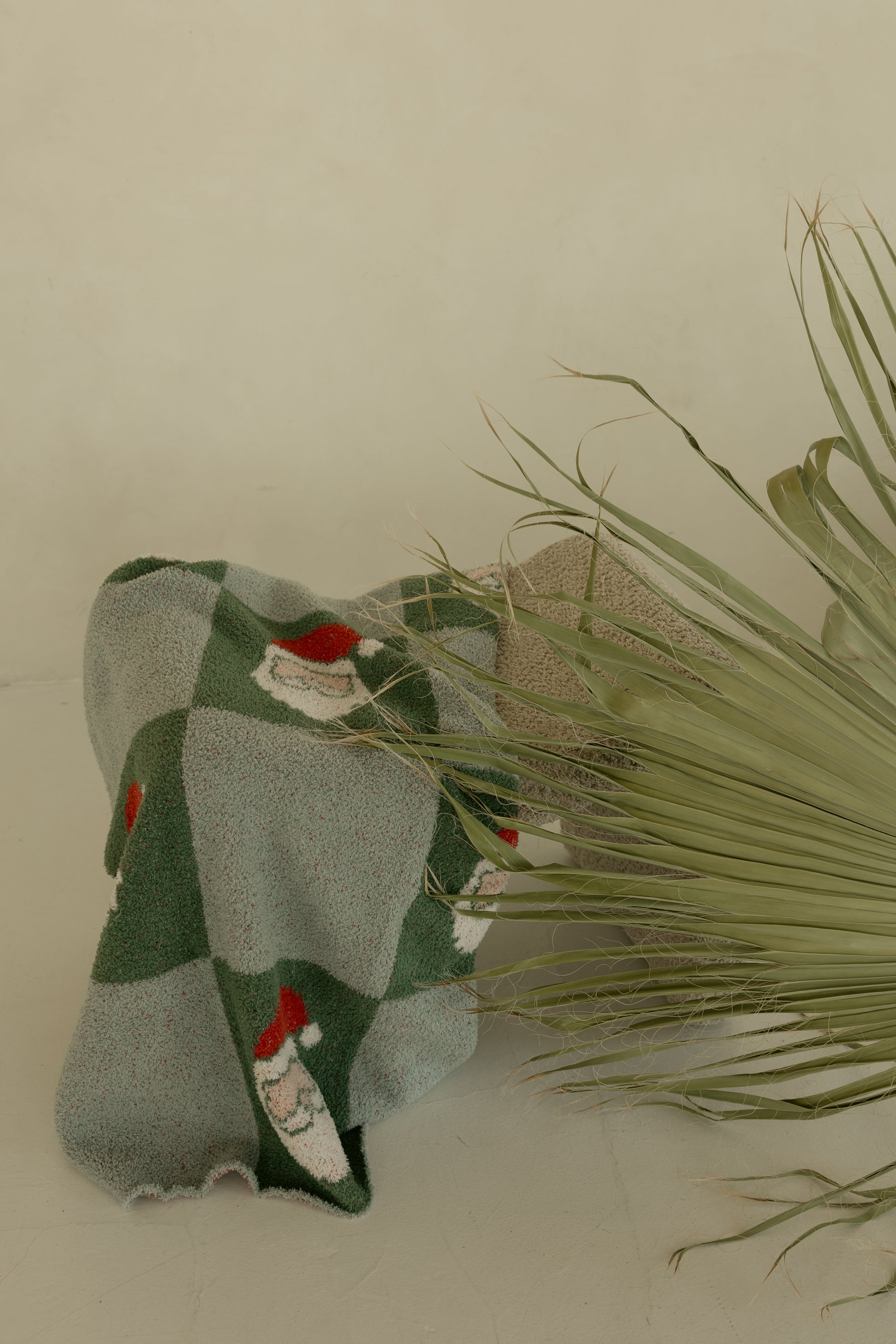 The Plush Blanket | Santa Checkerboard by forever french baby, featuring green and gray checks with Santa faces, is draped near a dried palm leaf against a beige wall.