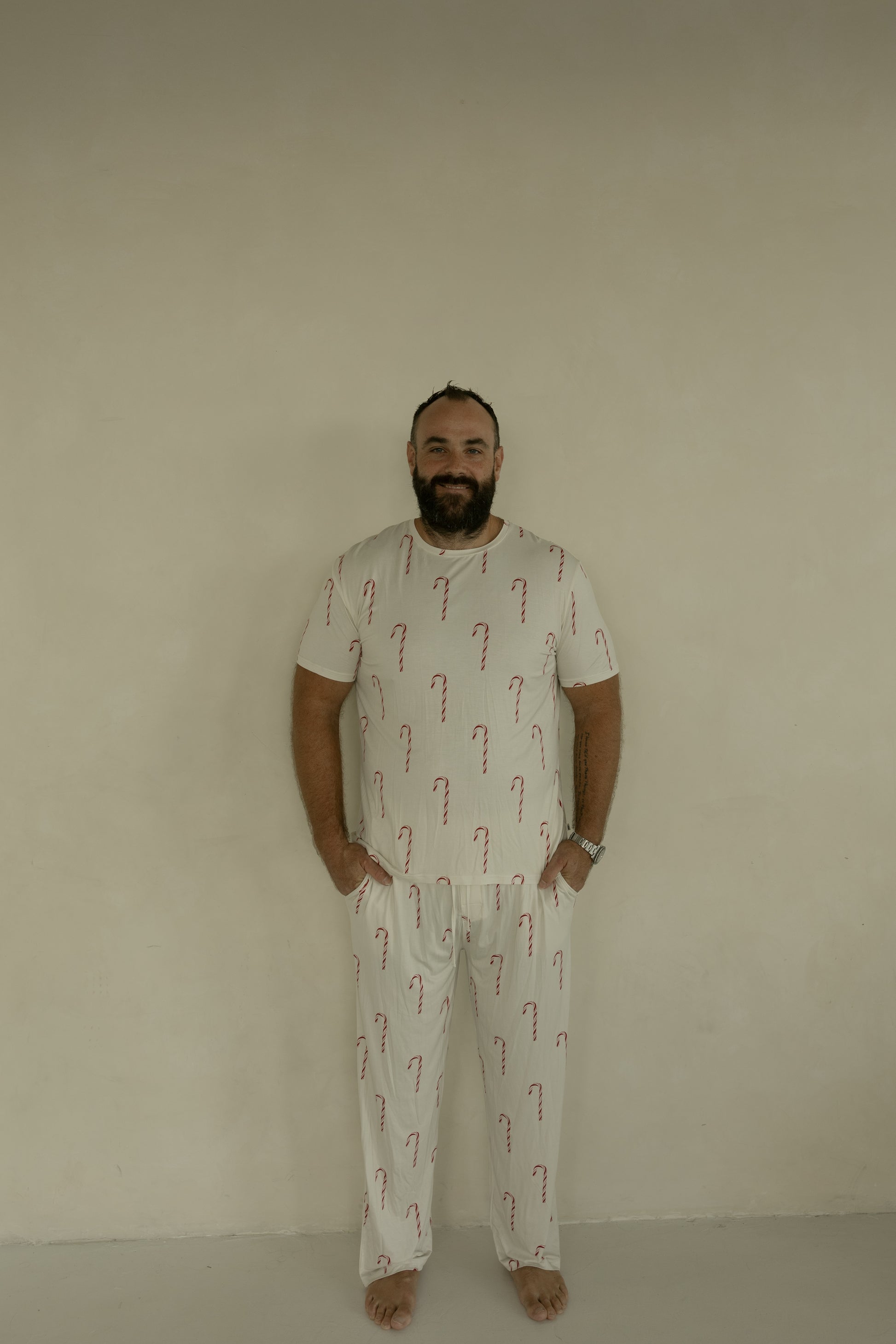A barefoot man stands against a light-colored wall, smiling with his hands in his pockets. He wears forever french baby's Men's Short Sleeve Bamboo Pajamas | Candy Cane, highlighting the comfort and style of these hypoallergenic PJs.