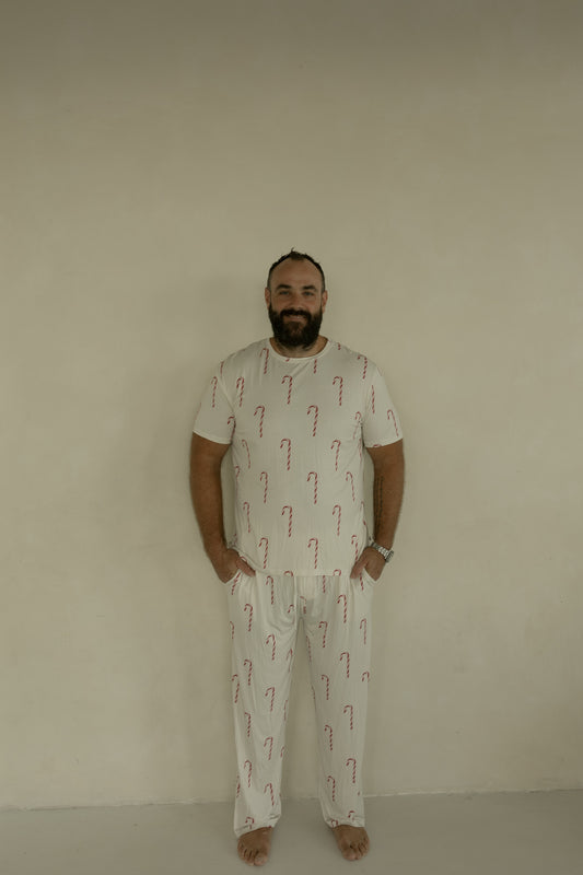 A barefoot man stands against a light-colored wall, smiling with his hands in his pockets. He wears forever french baby's Men's Short Sleeve Bamboo Pajamas | Candy Cane, highlighting the comfort and style of these hypoallergenic PJs.