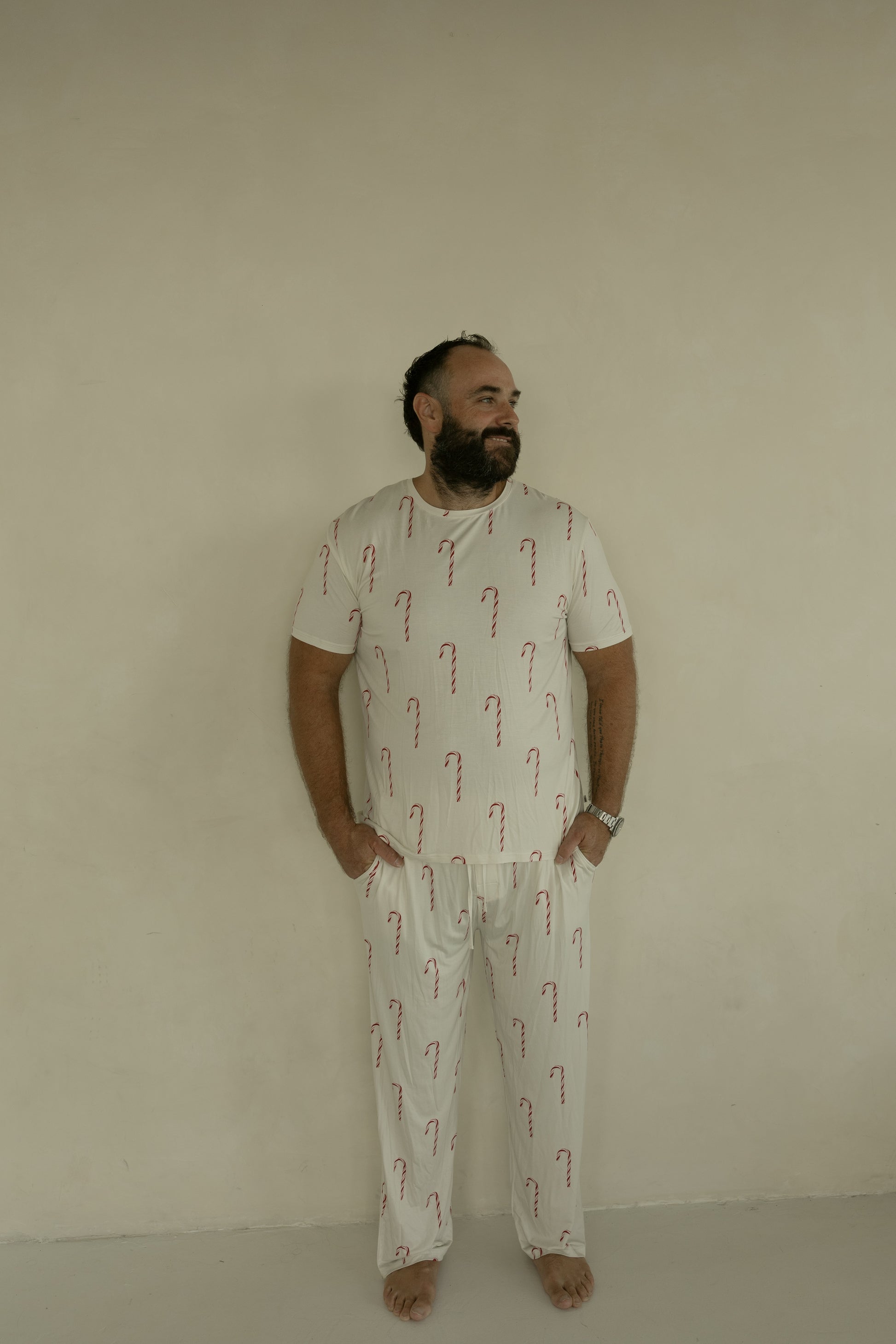 A bearded man stands barefoot by a beige wall, hands in pockets, wearing forever french baby’s Men's Short Sleeve Bamboo Pajamas | Candy Cane, and enjoys the comfort of bamboo sleepwear.
