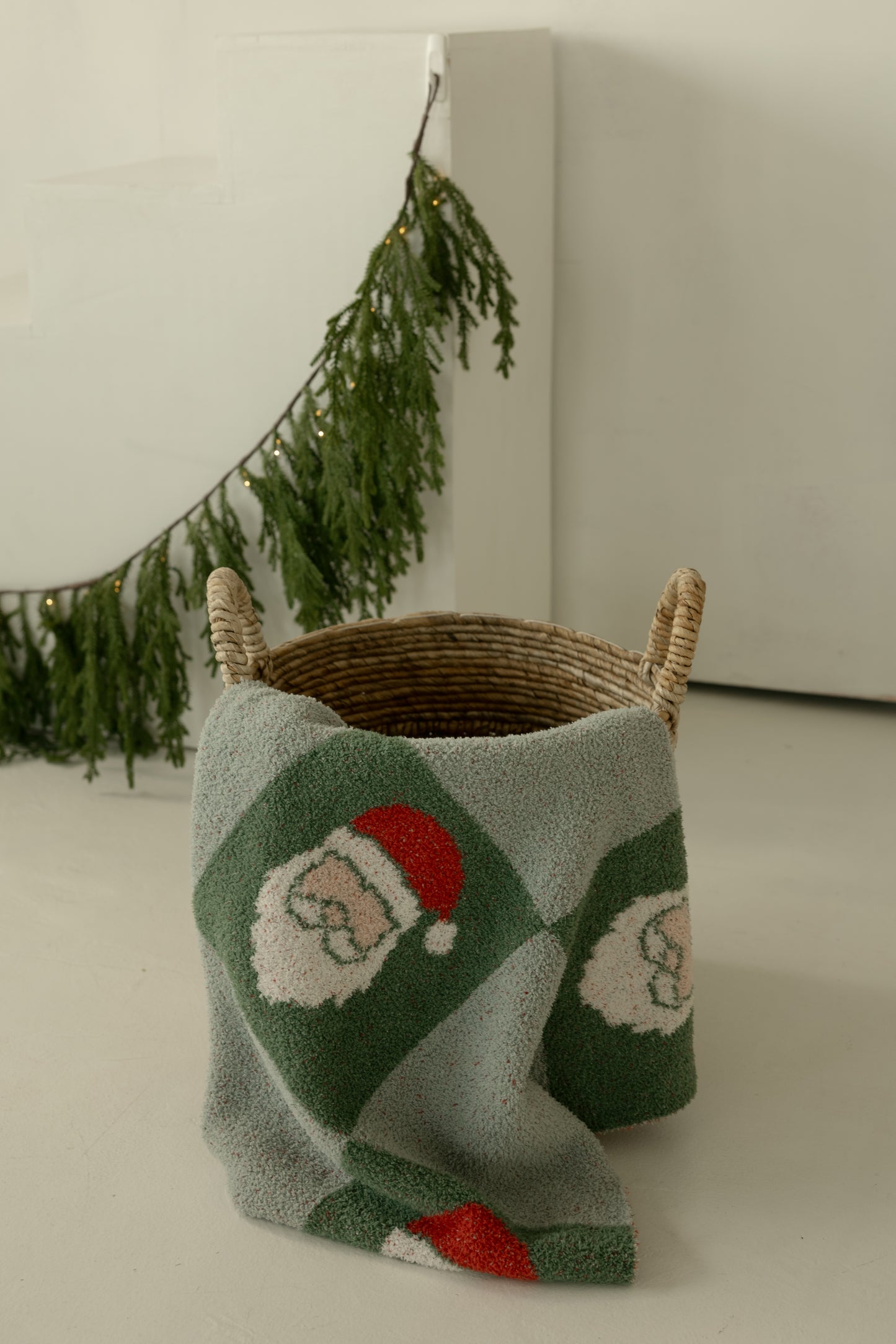 A woven basket with two handles sits on a light floor, draped with the forever french baby Plush Blanket | Santa Checkerboard. Fir garland hangs in the background on a white wall.