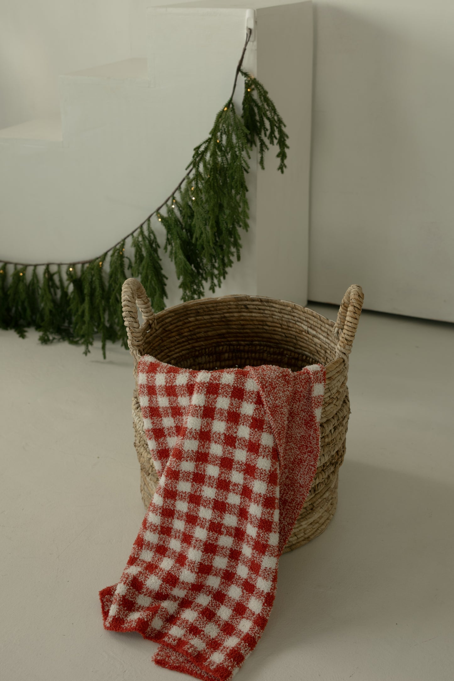 A woven basket with handles holds the forever French baby Plush Blanket | Red Gingham. In the background, a green garland with small lights decorates white stairs and a wall.