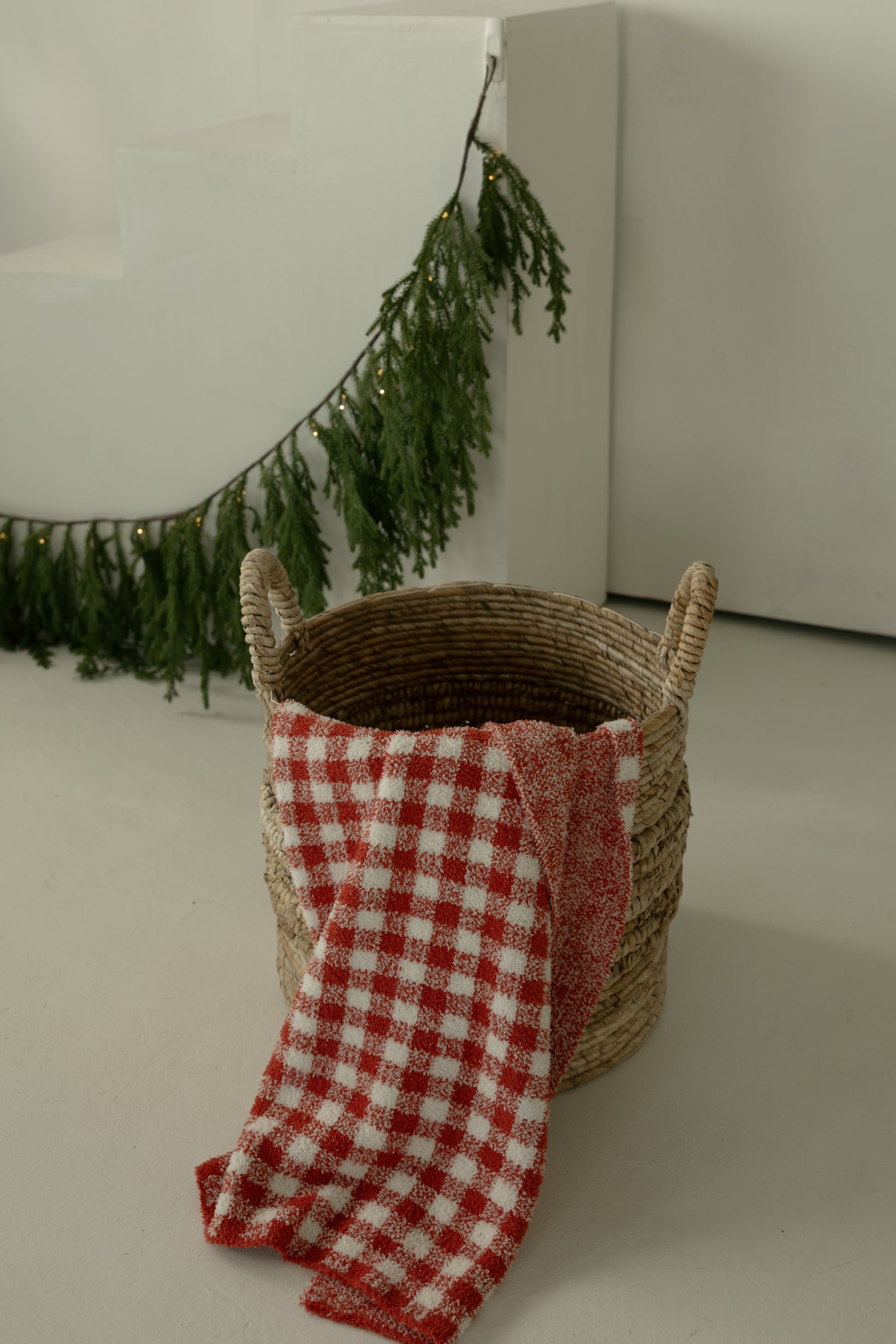 A woven basket with handles holds the forever French baby Plush Blanket | Red Gingham. In the background, a green garland with small lights decorates white stairs and a wall.