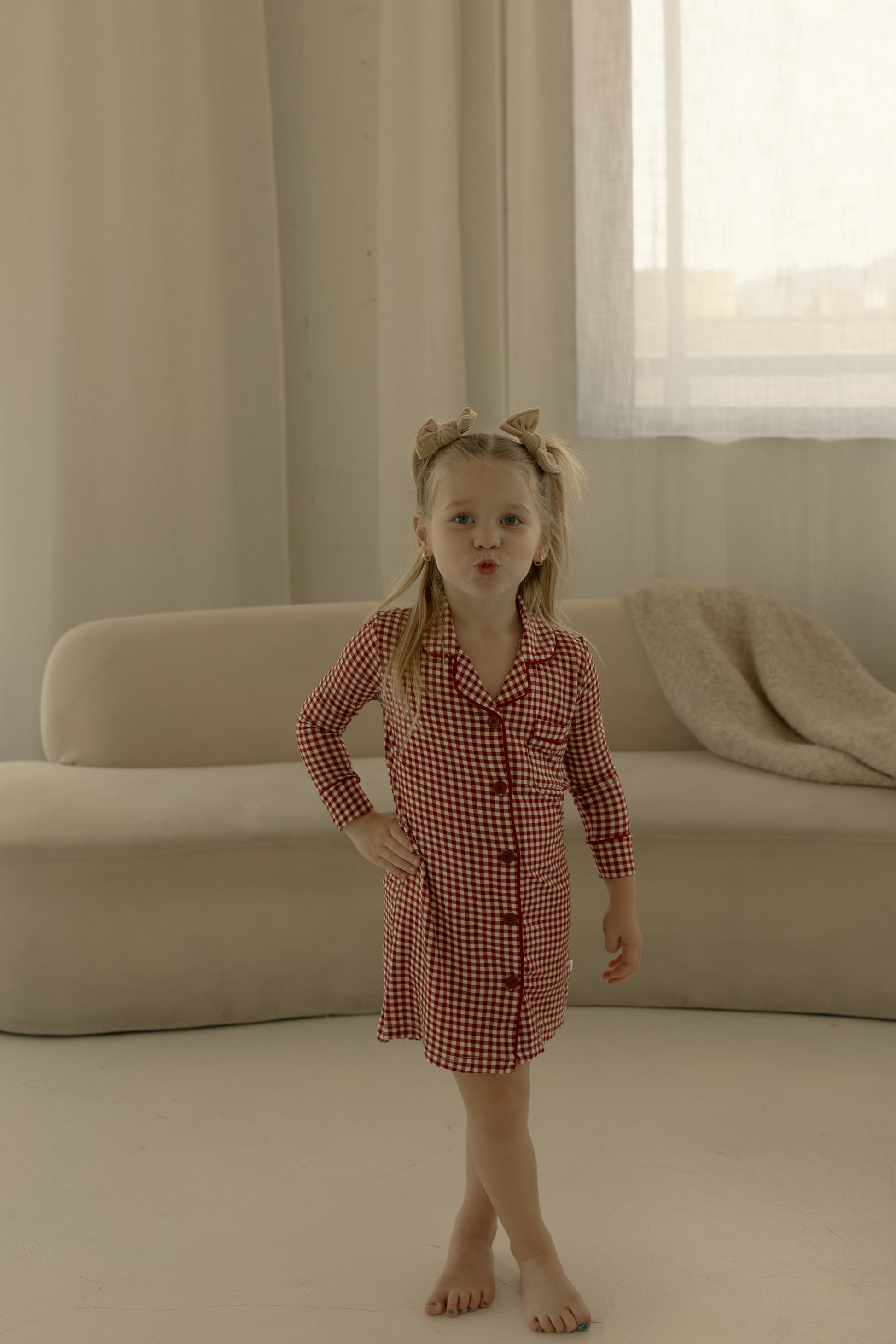 A young girl wears the forever french baby Toddler Bamboo Sleeping Dress in Red Gingham, barefoot indoors with a bow headband; behind her are a beige sofa and blanket.