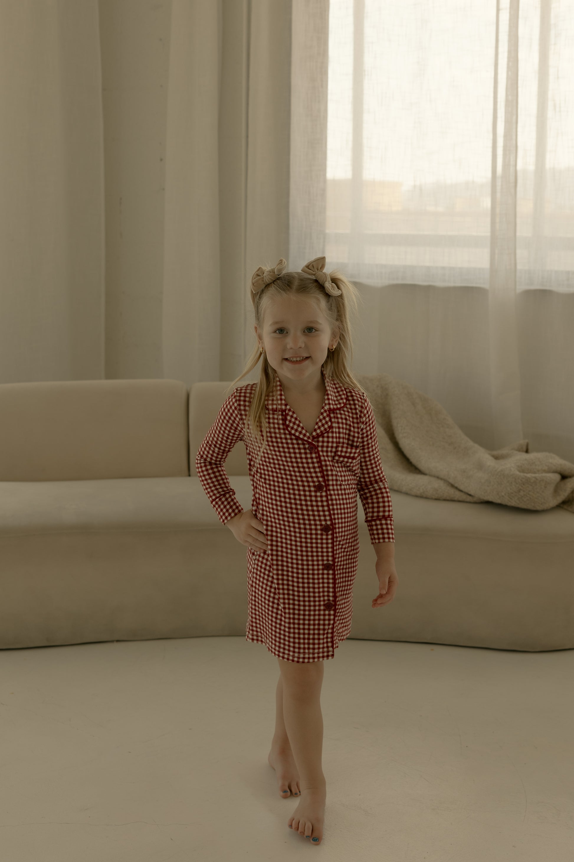 A young girl stands barefoot on a white floor wearing the forever french baby Toddler Bamboo Sleeping Dress in Red Gingham, with a bow in her hair, in front of a beige sofa draped with a blanket and sheer curtains behind her.