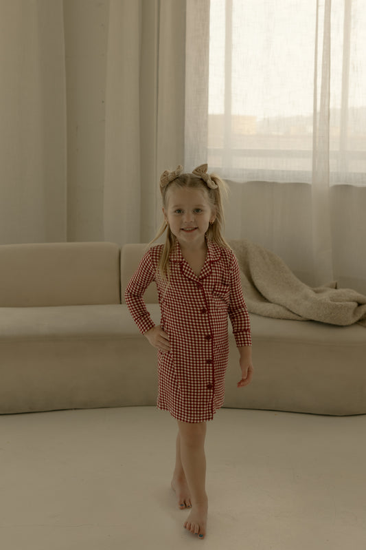 A young girl stands barefoot on a white floor wearing the forever french baby Toddler Bamboo Sleeping Dress in Red Gingham, with a bow in her hair, in front of a beige sofa draped with a blanket and sheer curtains behind her.