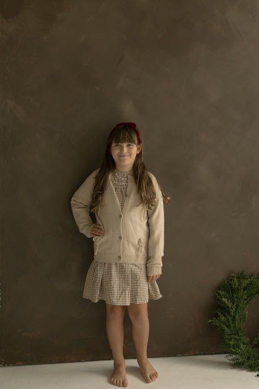 A young girl with long brown hair and a red headband stands barefoot against a brown wall, wearing the forever french baby Knit Cardigan | Bows over a checked dress. She smiles softly with one hand on her hip, greenery in the bottom right corner.