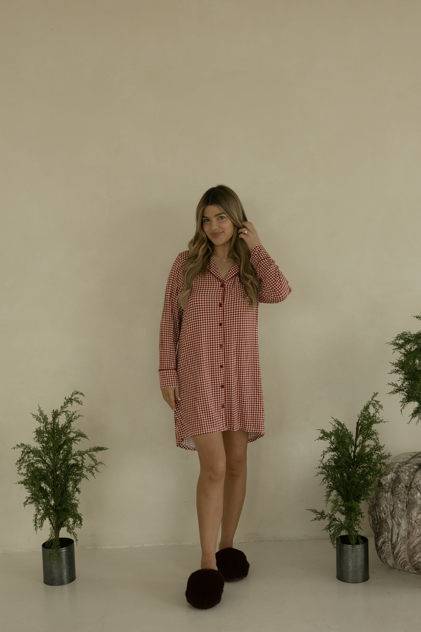 A woman with long wavy hair smiles indoors, wearing the forever french baby Women's Bamboo Sleeping Dress in Red Gingham, paired with dark slippers and surrounded by green plants—effortless style and comfort in bamboo sleepwear.