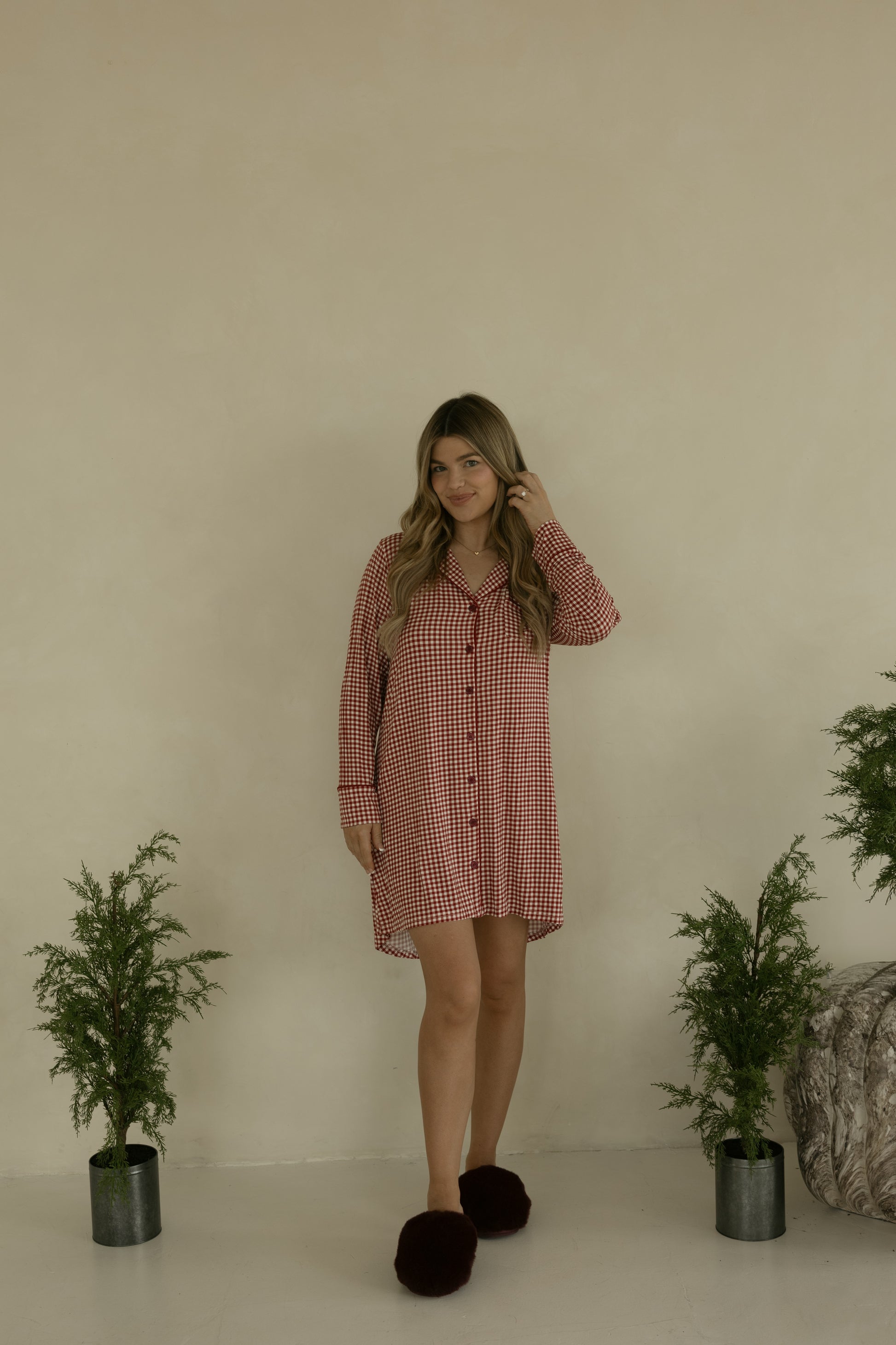 A woman with long wavy hair smiles indoors, wearing the forever french baby Women's Bamboo Sleeping Dress in Red Gingham, paired with dark slippers and surrounded by green plants—effortless style and comfort in bamboo sleepwear.