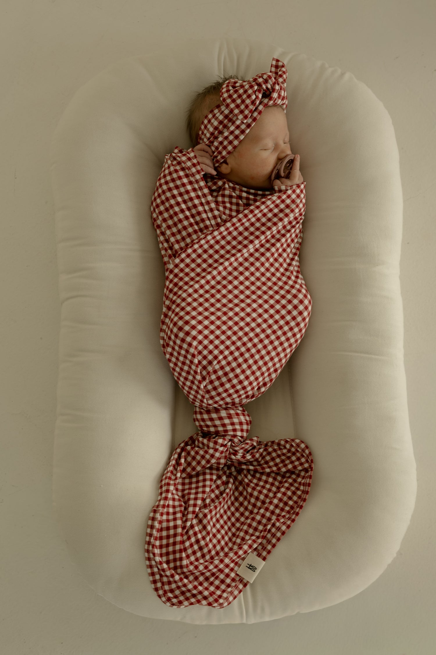 A newborn sleeps peacefully on a soft white cushion, swaddled in the forever french baby Bamboo Swaddle in Red Gingham with a matching headband.