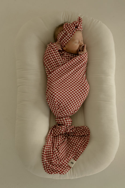 A newborn sleeps peacefully on a soft white cushion, swaddled in the forever french baby Bamboo Swaddle in Red Gingham with a matching headband.