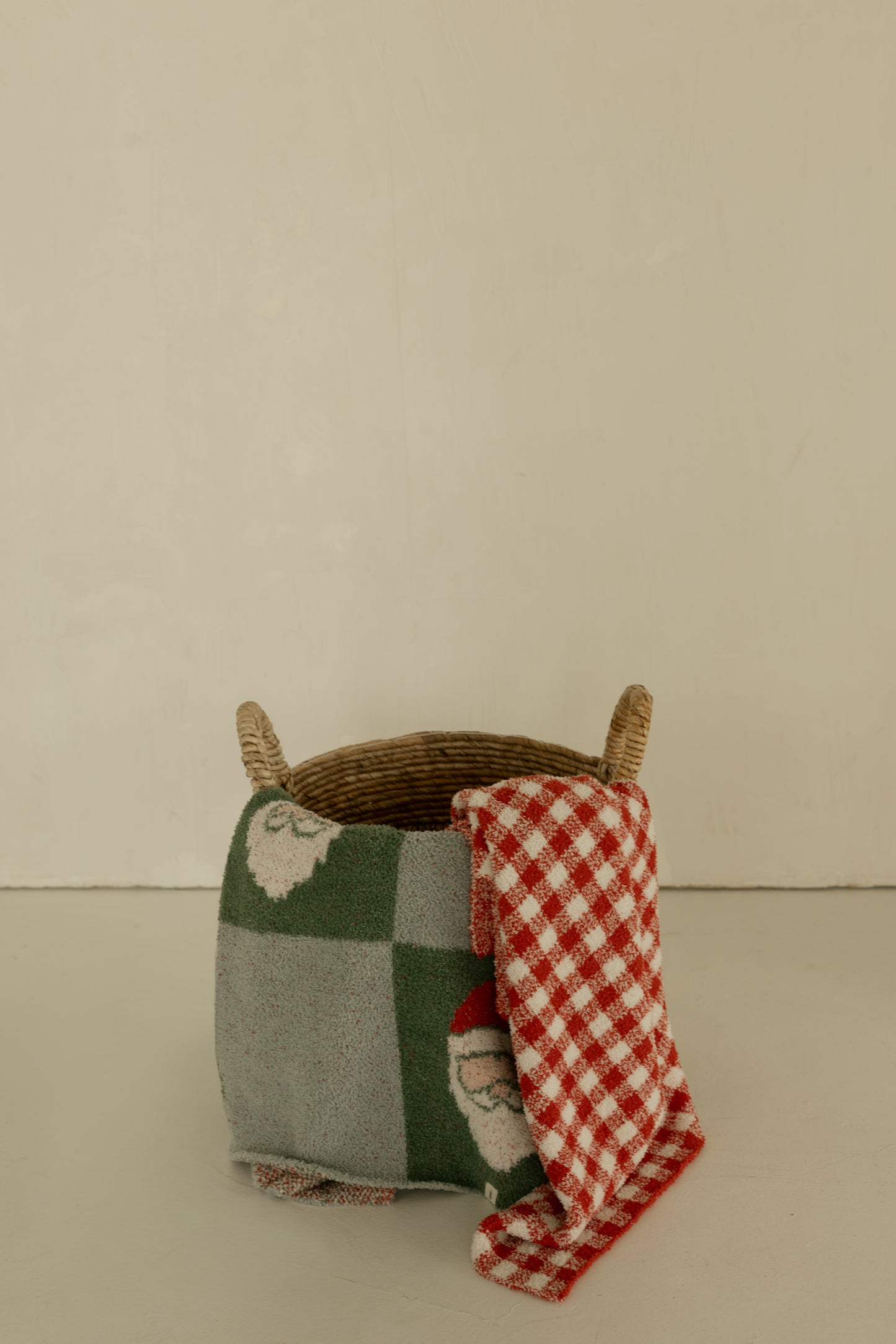 A wicker basket with handles is topped with the forever French baby Plush Blanket | Red Gingham, featuring Santa faces and red-and-white checks, set against a plain beige background.