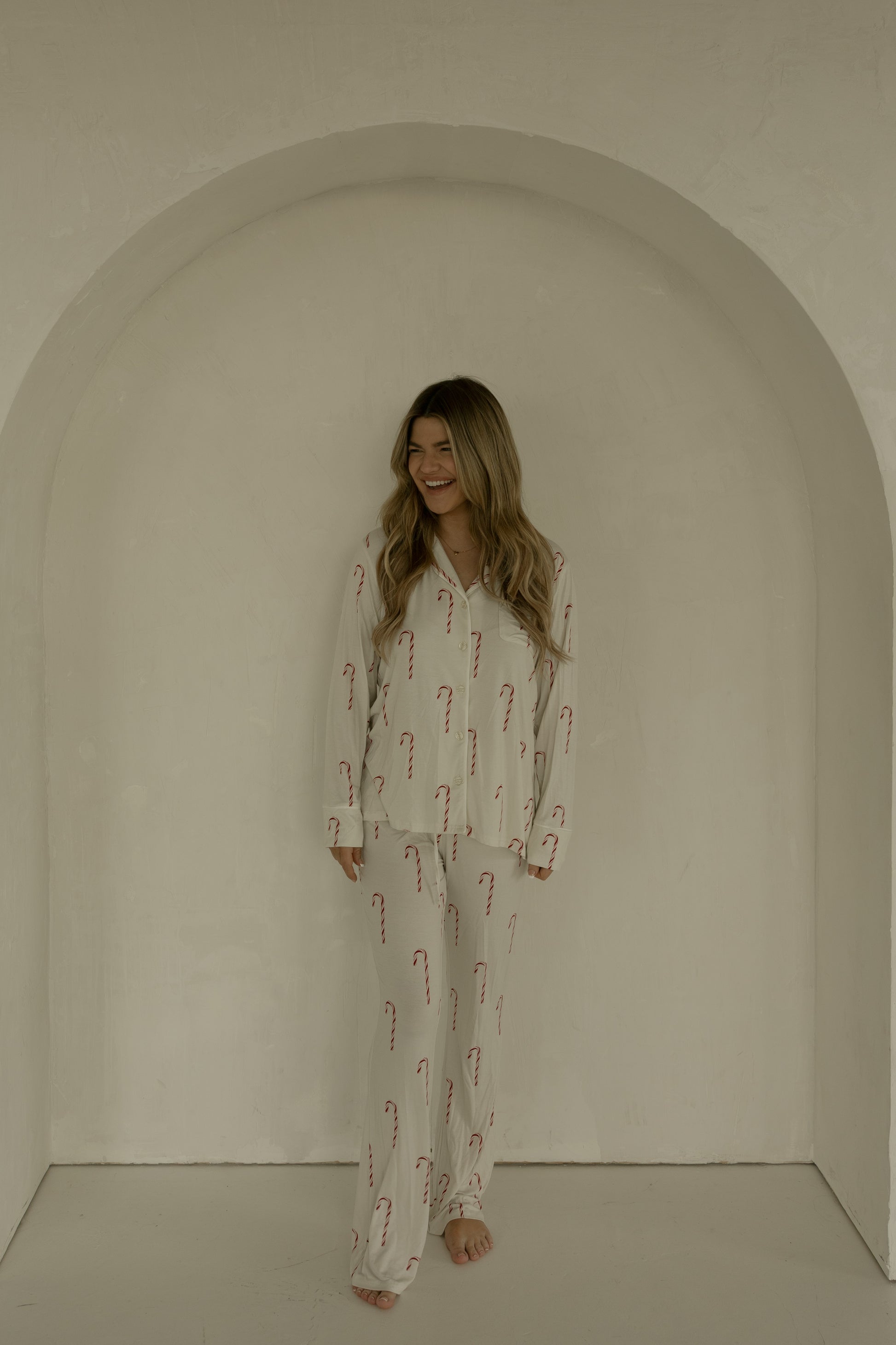 A woman with long, wavy hair stands barefoot in a minimal, light archway, smiling and wearing forever french baby’s Long Sleeve Women's Bamboo Pajamas | Candy Cane—a cream set with red candy cane patterns.