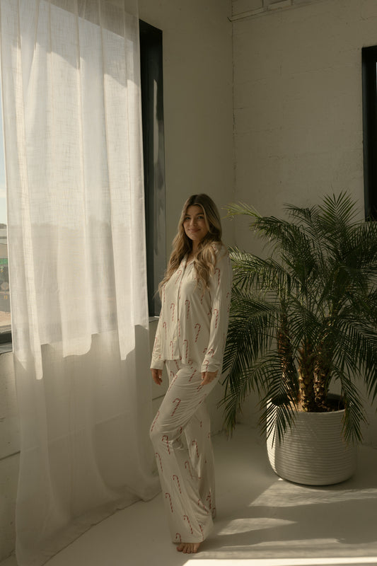 A woman stands barefoot in a sunlit room near a palm plant and sheer curtains, wearing forever french baby's Long Sleeve Women's Bamboo Pajamas in the Candy Cane pattern.