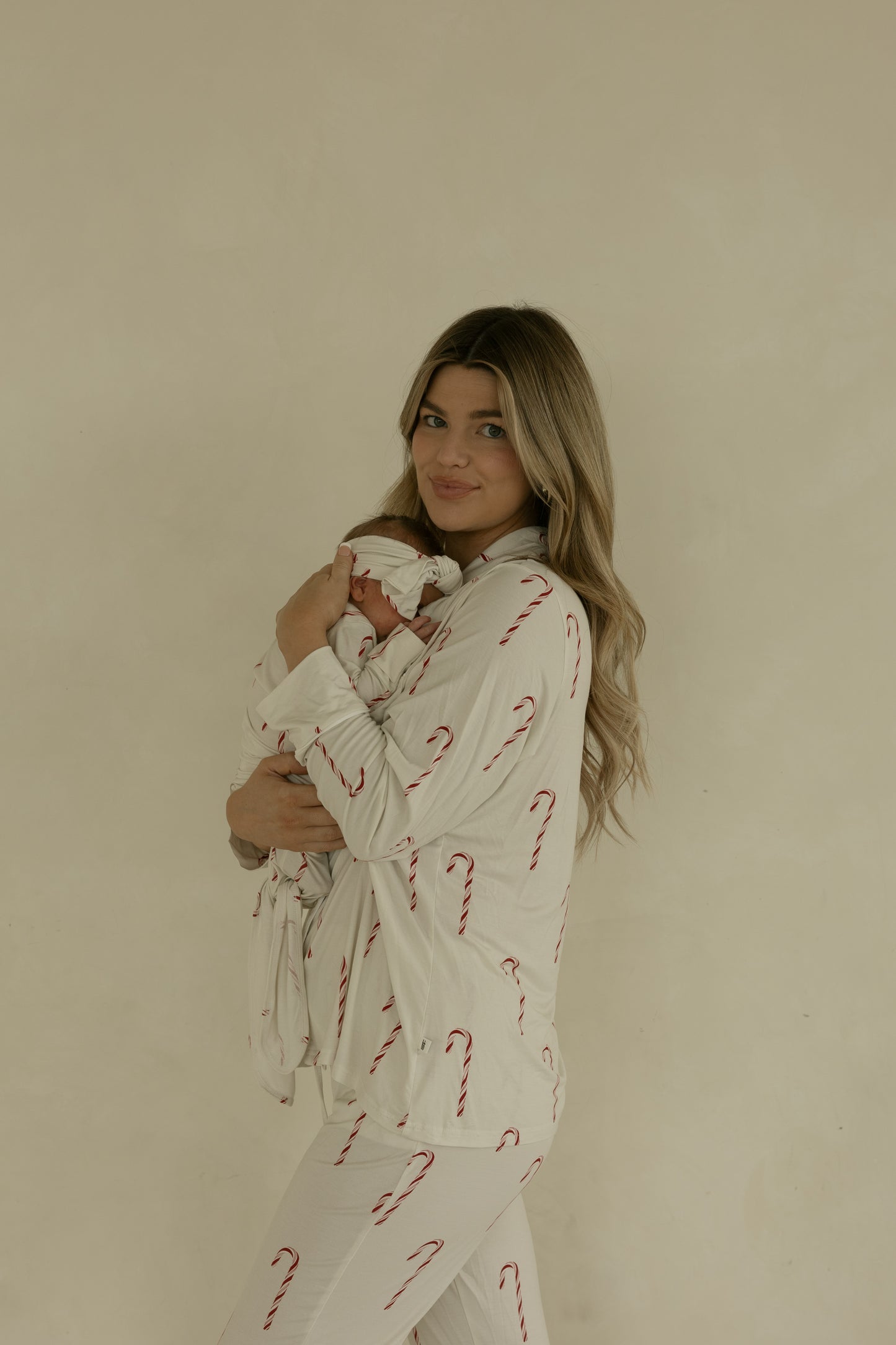 A woman with long blonde hair holds a swaddled newborn, both in matching forever french baby Long Sleeve Women's Bamboo Pajamas | Candy Cane. The white pajamas feature red candy cane prints and are set against a plain beige background.