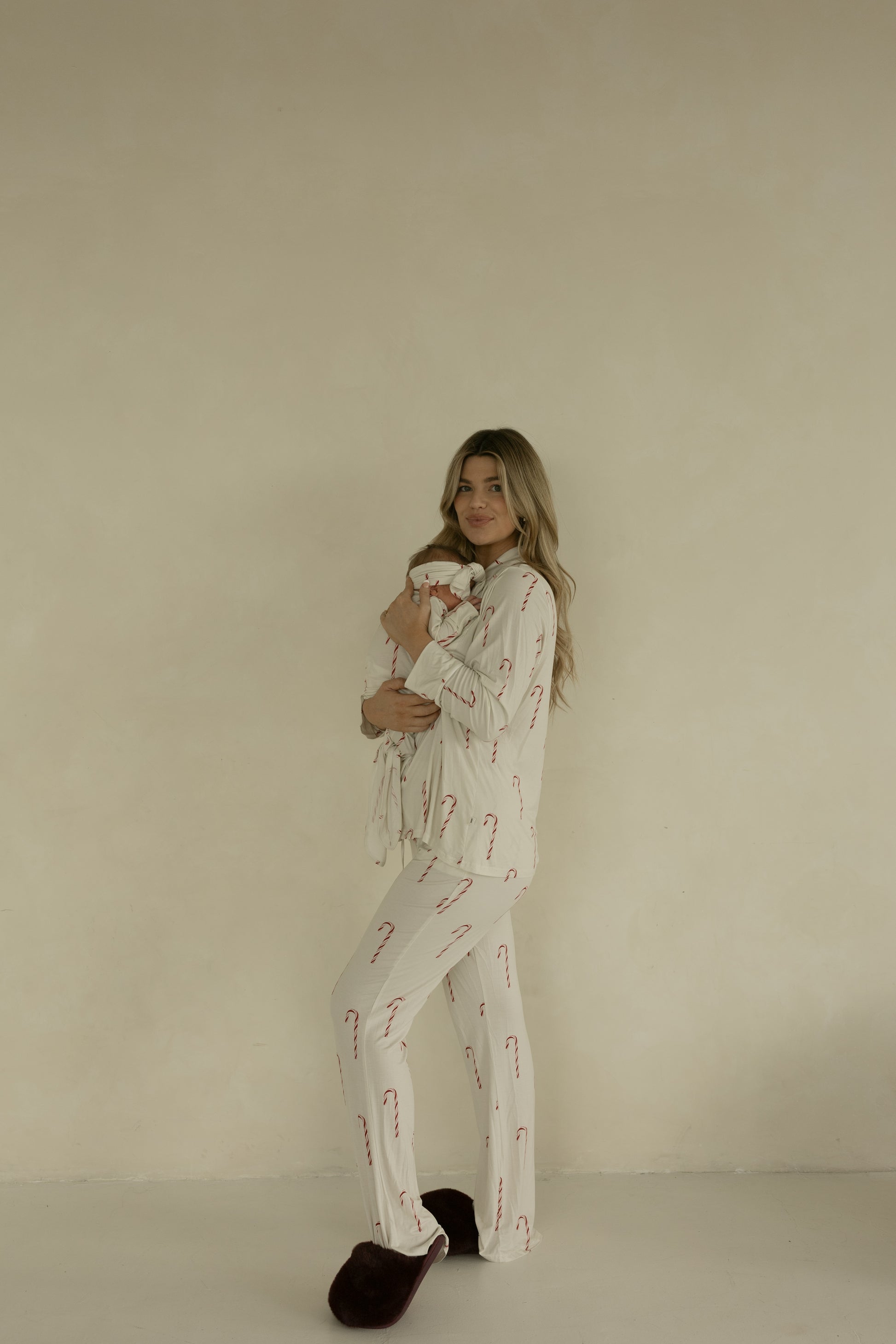 A woman wears forever french baby’s Long Sleeve Women's Bamboo Pajamas | Candy Cane—cream with red candy cane prints—standing against a beige wall, holding a baby in white, and smiling at the camera in dark slippers.