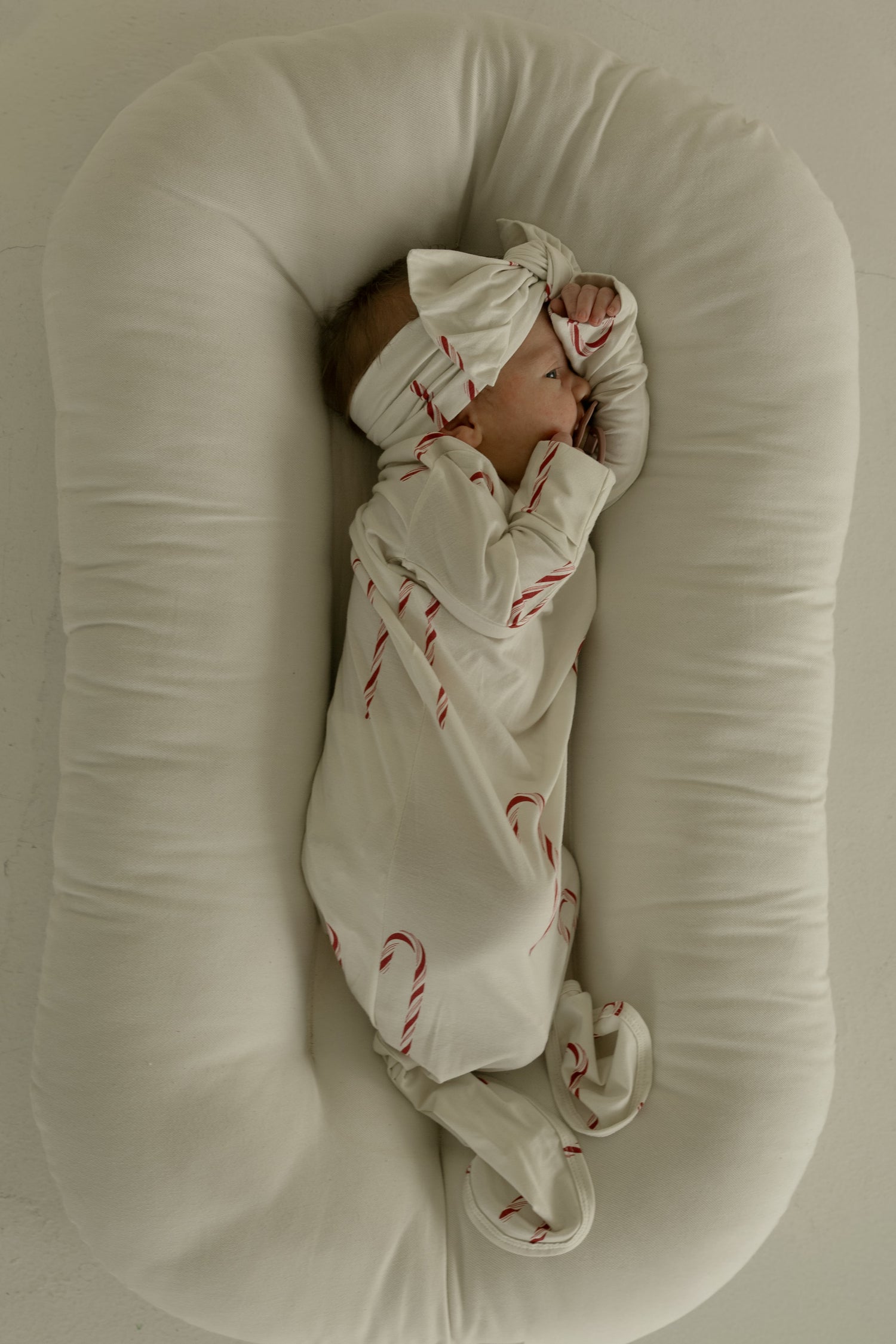 A baby in the forever french baby Bamboo Knotted Gown | Candy Cane—cream-colored with red candy cane patterns and a matching headband—lies sideways on a soft, white oval cushion with hands near their face.