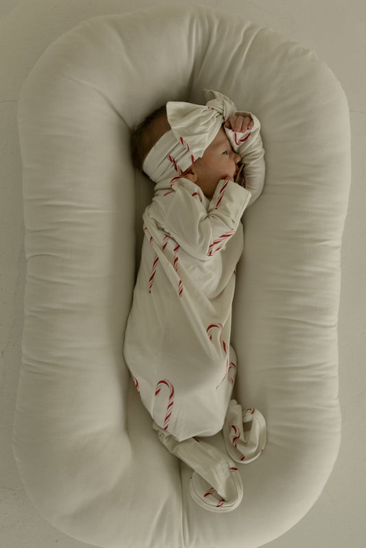 A baby in the forever french baby Bamboo Knotted Gown | Candy Cane—cream-colored with red candy cane patterns and a matching headband—lies sideways on a soft, white oval cushion with hands near their face.