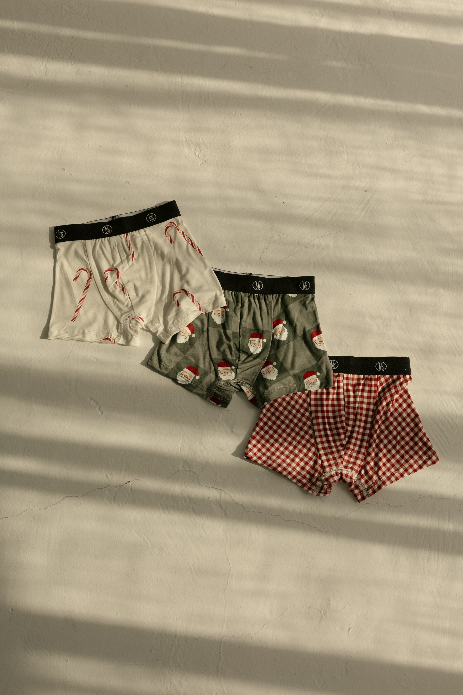 The forever french baby Bamboo Boxers (3 Pack) set features festive men’s boxers in red gingham, white with candy canes, and green with Santa checkerboard designs, laid out on a light surface with striped shadows.
