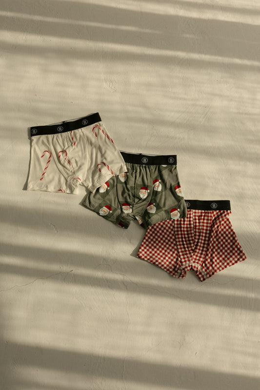 The forever french baby Bamboo Boxers (3 Pack) set features festive men’s boxers in red gingham, white with candy canes, and green with Santa checkerboard designs, laid out on a light surface with striped shadows.