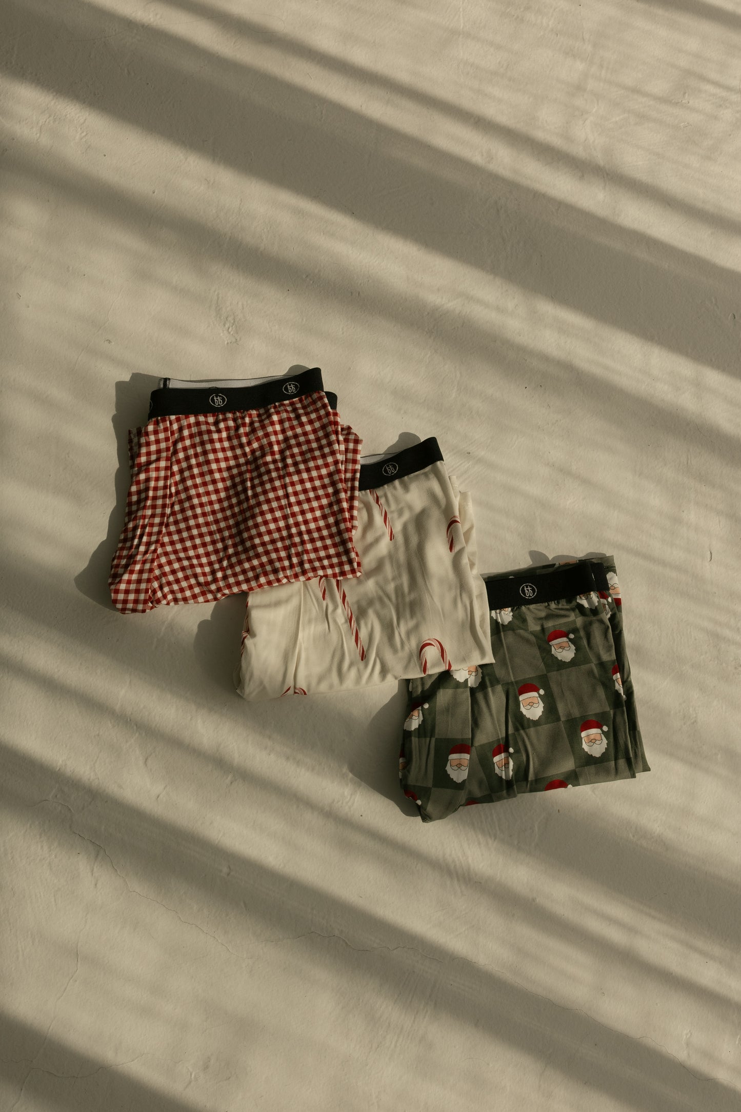 Three pairs of forever french baby Men's Bamboo Boxers (Red Gingham, Candy Cane & Santa Checkerboard) are folded on a sunlit surface, shadows from window blinds stretching across these comfy underwear.