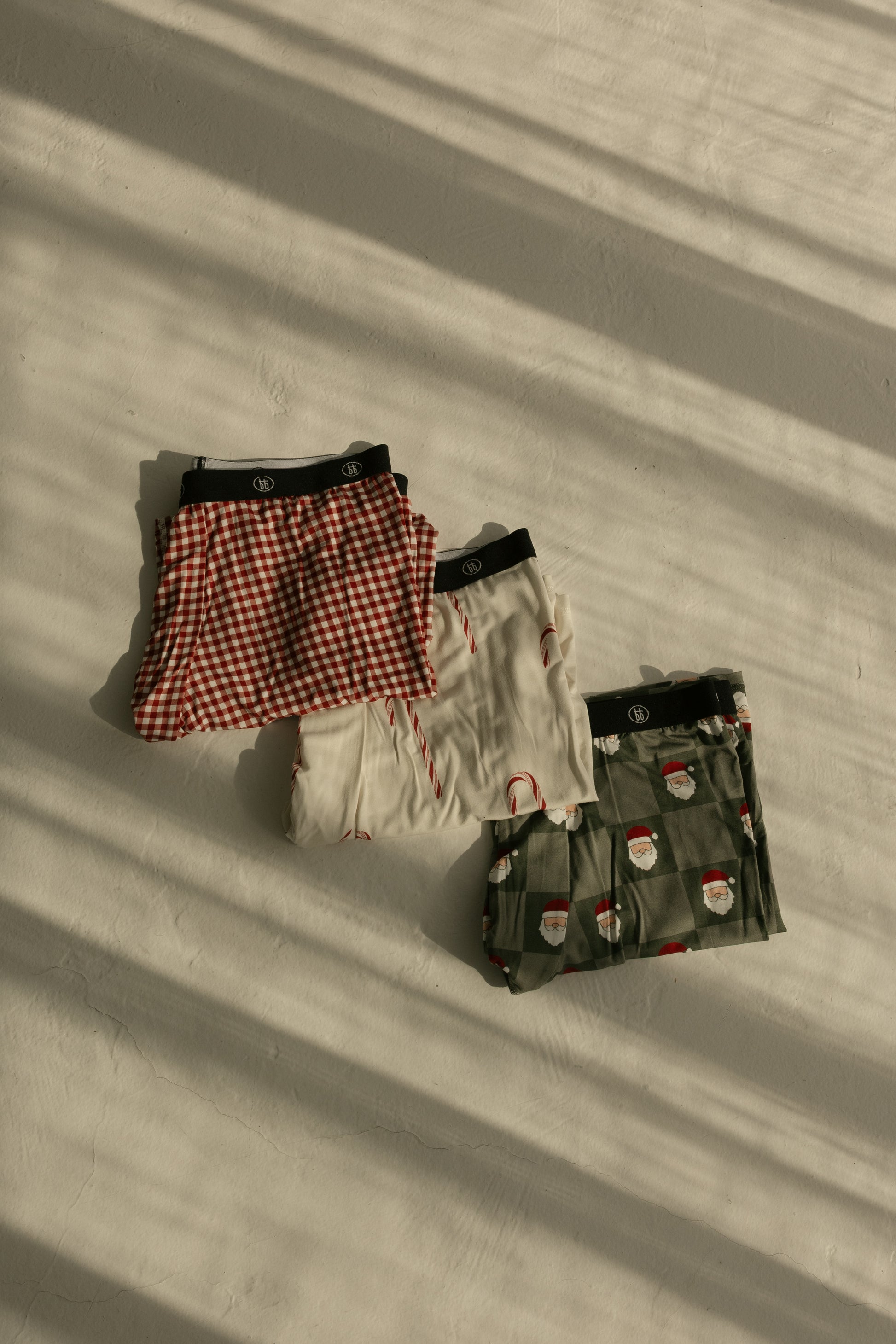 Three pairs of forever french baby Men's Bamboo Boxers (Red Gingham, Candy Cane & Santa Checkerboard) are folded on a sunlit surface, shadows from window blinds stretching across these comfy underwear.