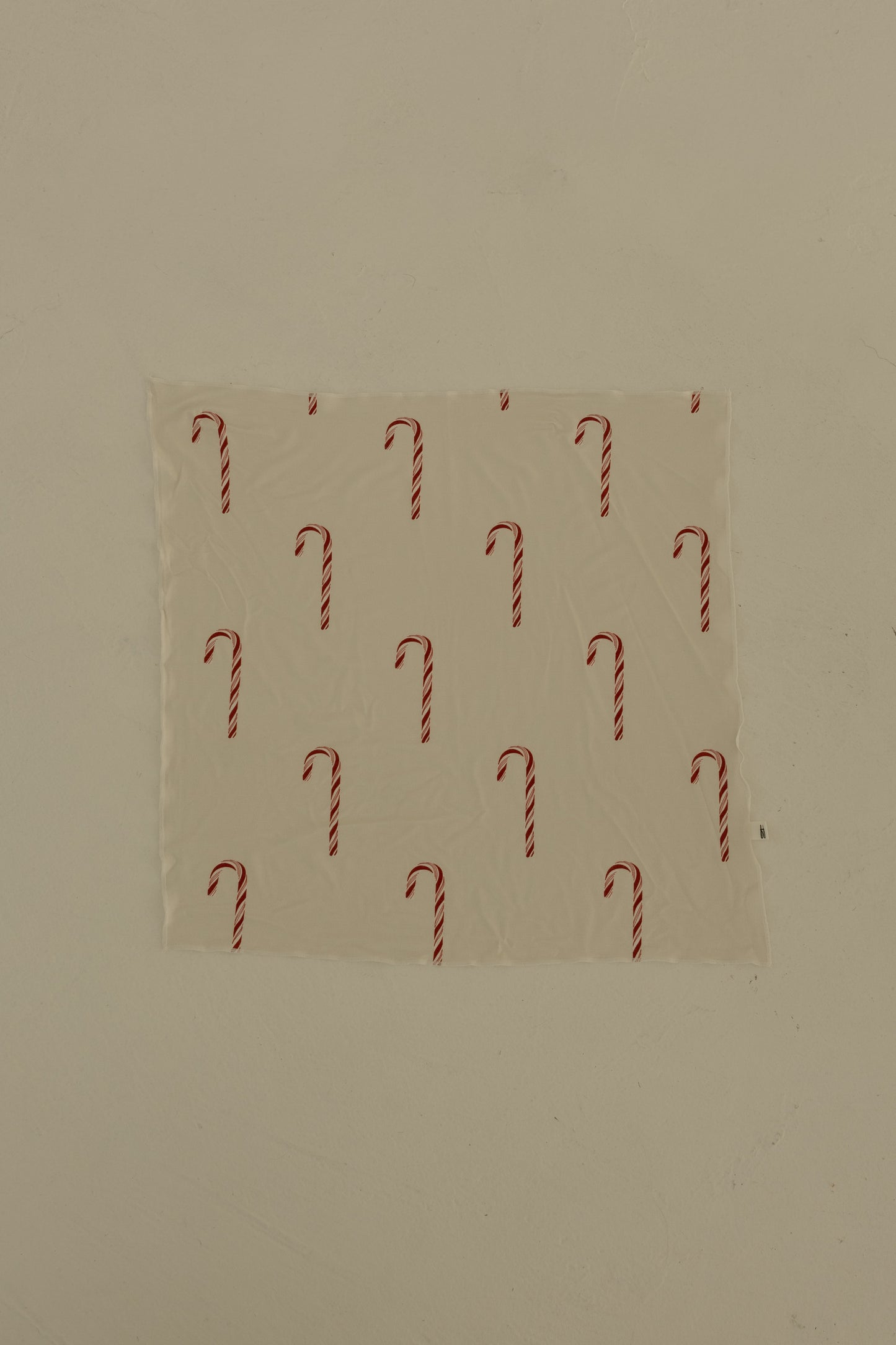 The Bamboo Bandana | Candy Cane by forever french baby, featuring a repeating candy cane print, is taped to a plain beige wall.