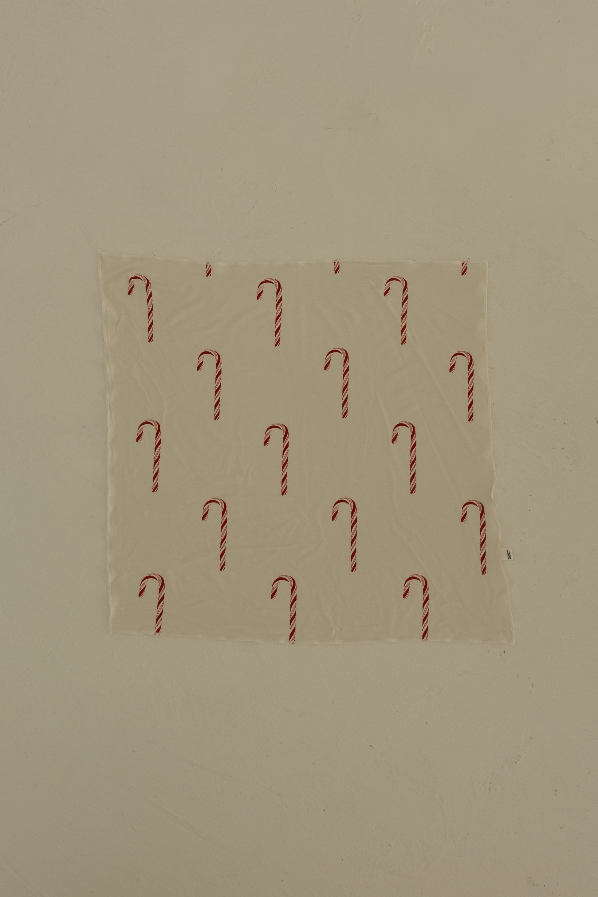 The Bamboo Bandana | Candy Cane by forever french baby, featuring a repeating candy cane print, is taped to a plain beige wall.